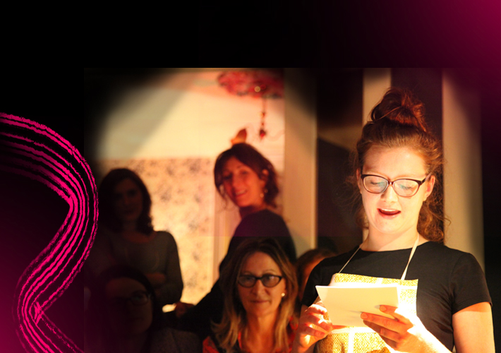 Artist Michelle Hall is performing. They have long brown hair tied in a bun and black framed glasses. They are in a room full of people listening attentively. The lights in the room have a warm orange glow. The left of the image fades to black and against this are bright cerise pink painterly lines.