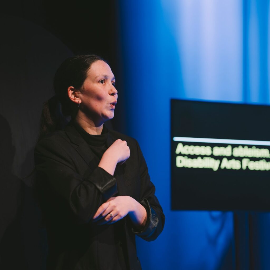 An ISL interpreter on the Disrupt festival stage. They gesture towards an audience. Behind them a screen shows captions. Photo by Leanne Sullivan.