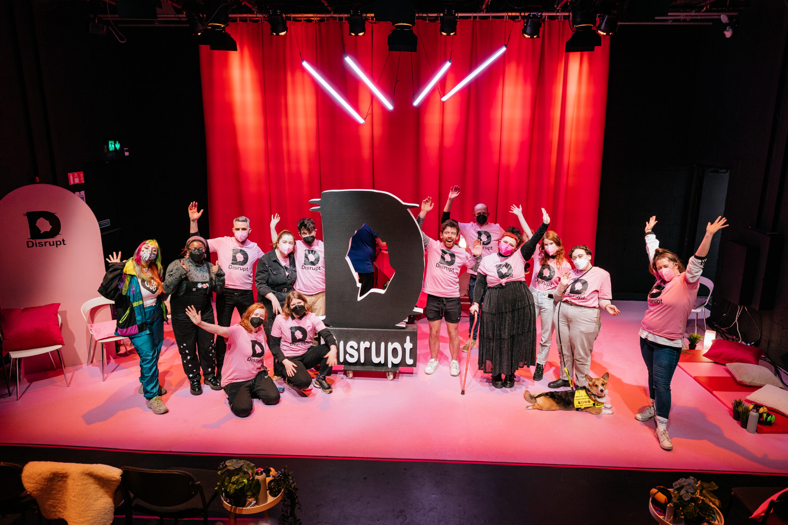 A group photo of 13 members of the Disrupt festival team on stage. They are wearing pink festival t-shirts printed with the Disrupt Logo. The stage is heavily clad in pink tones, a light pink carpet and hot pink curtains. They are also joined on stage by a corgi service dog and a giant foam cutout of the Disrupt Logo in black. Some strip lighting is hanging overhead. Photo by Simon Lazewski.