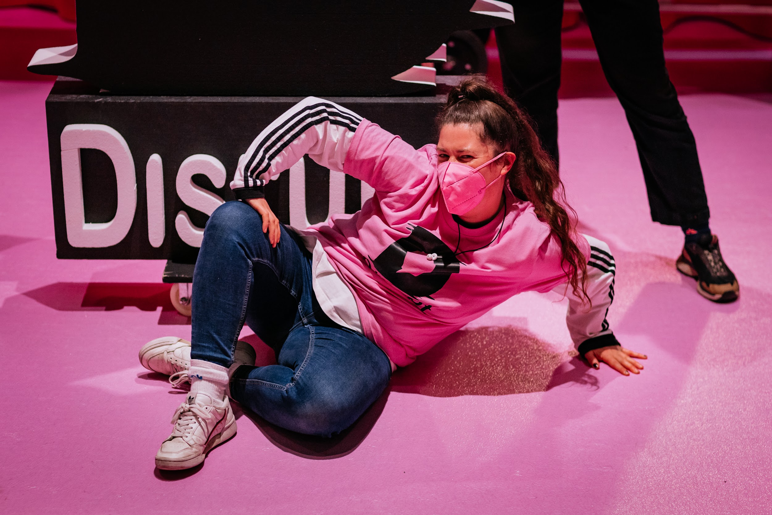 Festival crew member Ailbhe O’Connor poses for a photograph alongside Disrupt festival branding. She is lying on the ground, legs crossed and propped up on her arms. She is wearing a pink Disrupt t-shirt and pink facemask. She has a long pink ponytail and is winking to the camera.