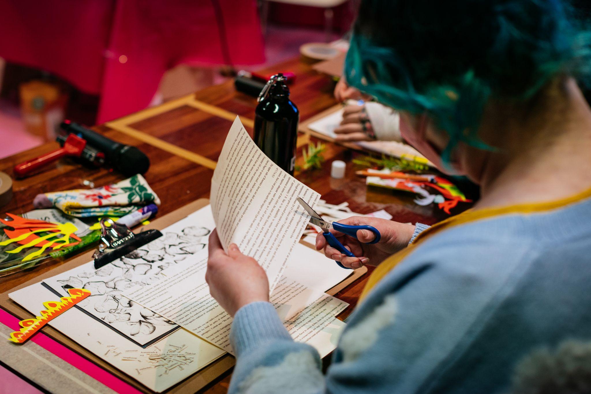 A person sitting at a desk, engaged in crafting activities. The person has long blue hair. They have fair skin, and wear a light cardigan and a yellow top. They appear to be cutting words from a page. On the table are various materials. These include texts, a clipboard, and paper in many colours. Photo by Simon Lazewski.