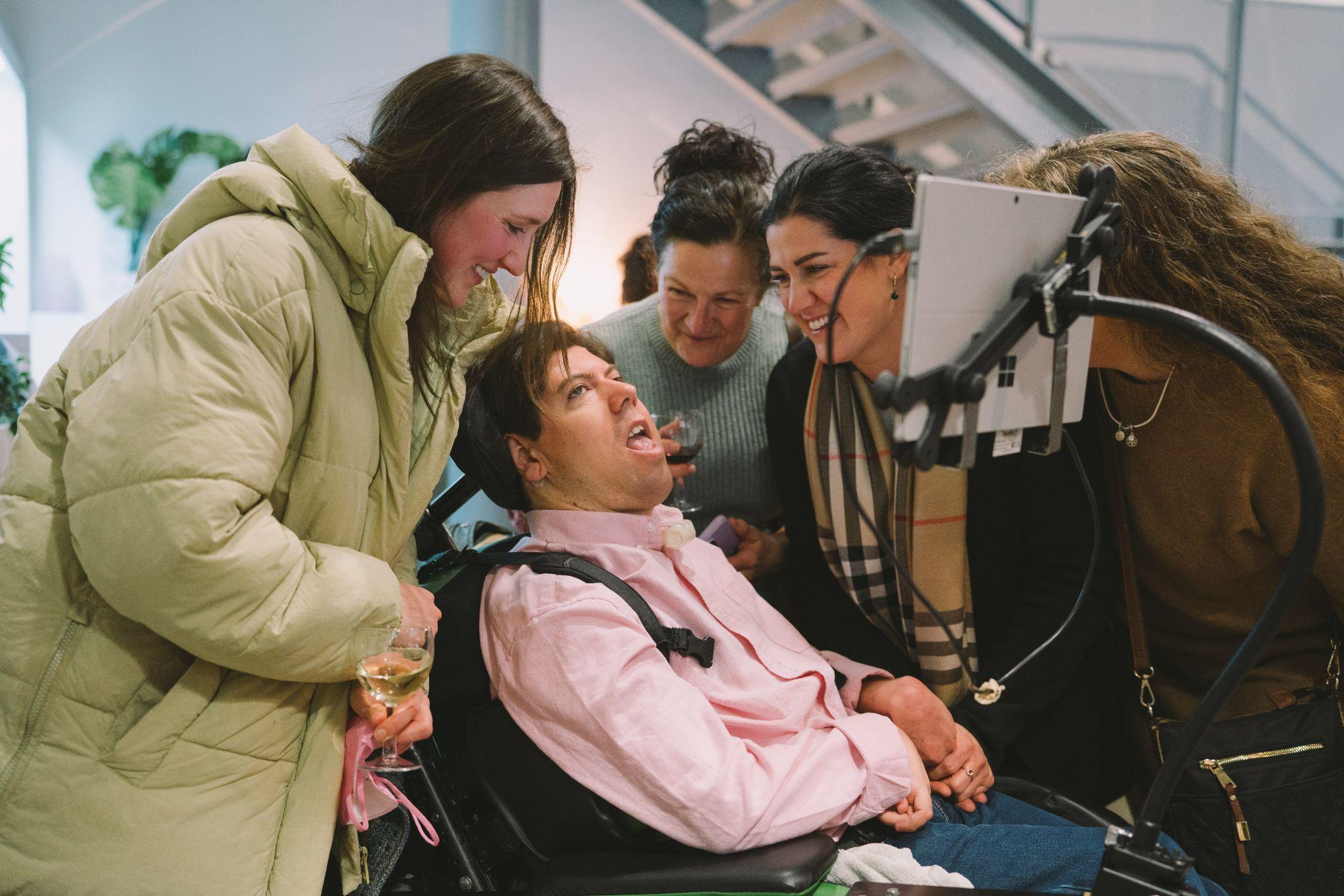 Artist David Parnell wearing a light pink shirt and blue trousers. David is seated in a wheelchair which has a screen attached to it. He is surrounded by a group of people with long dark hair. The group are smiling and appear excited. The background is out of focus but features a white room and a staircase. Photo by Leanne Sullivan.