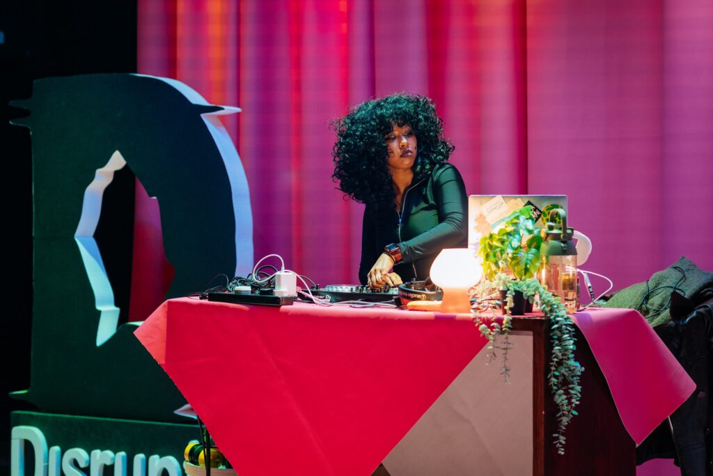 A table with plants, lights, a pink covering and DJ equipment is set up on the Disrupt stage. DJ Renn Miano plays a set. Renn has voluminous dark curls and is concentrated on their work. Renn wears a black lycra zip top. They are lit in pink, blue and green hues. Behind Renn is a pink curtain and a large styrofoam D, the disrupt logo. Photo by Simon Lazewski.