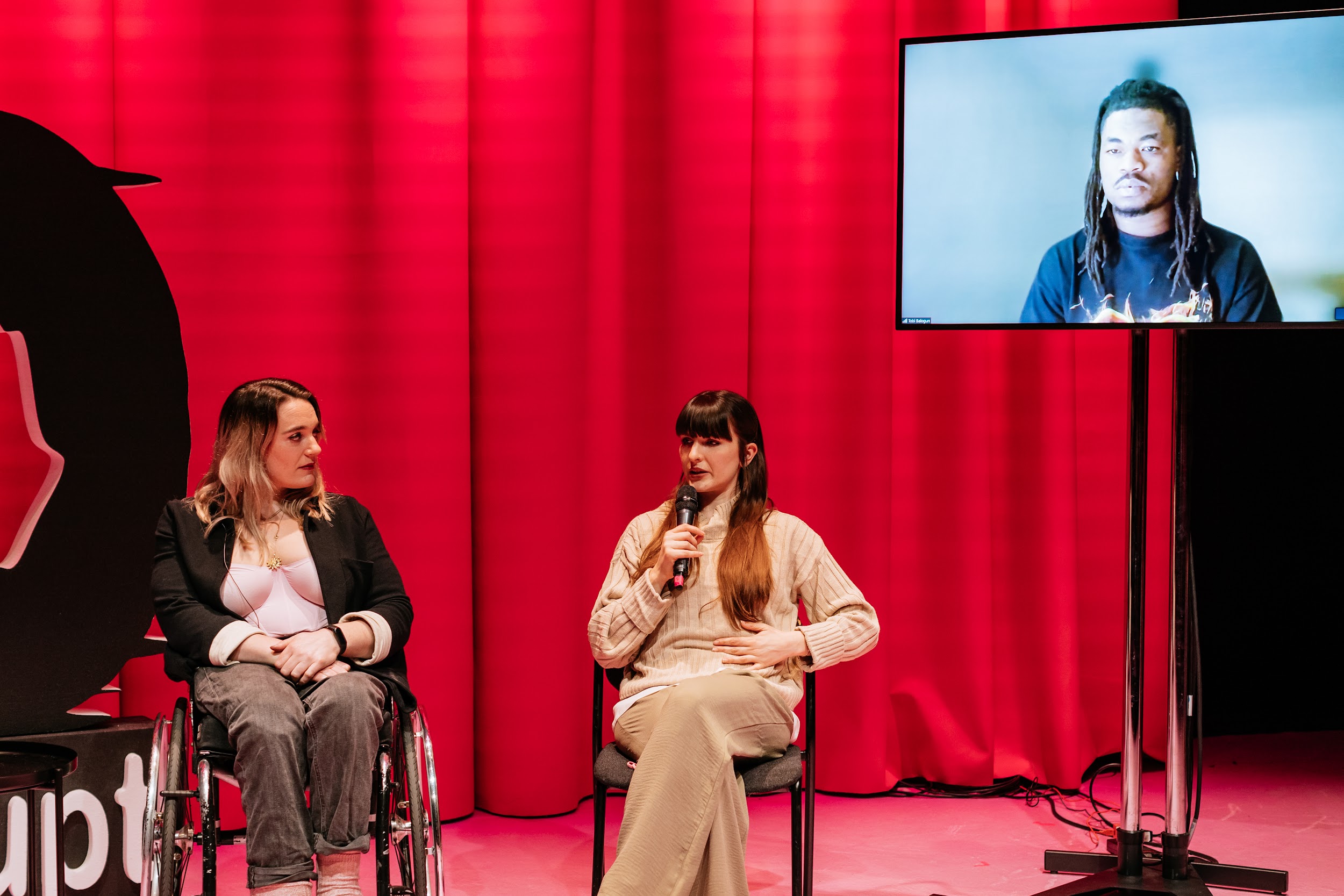 Three speakers at a Disrupt festival panel talk. Louise Bruton, a wheelchair user, looks to panellist Alison Clarke. Clarke speaks into a microphone. From a screen, Tobi Balogun looks on. Photo by Simon Lazewski.