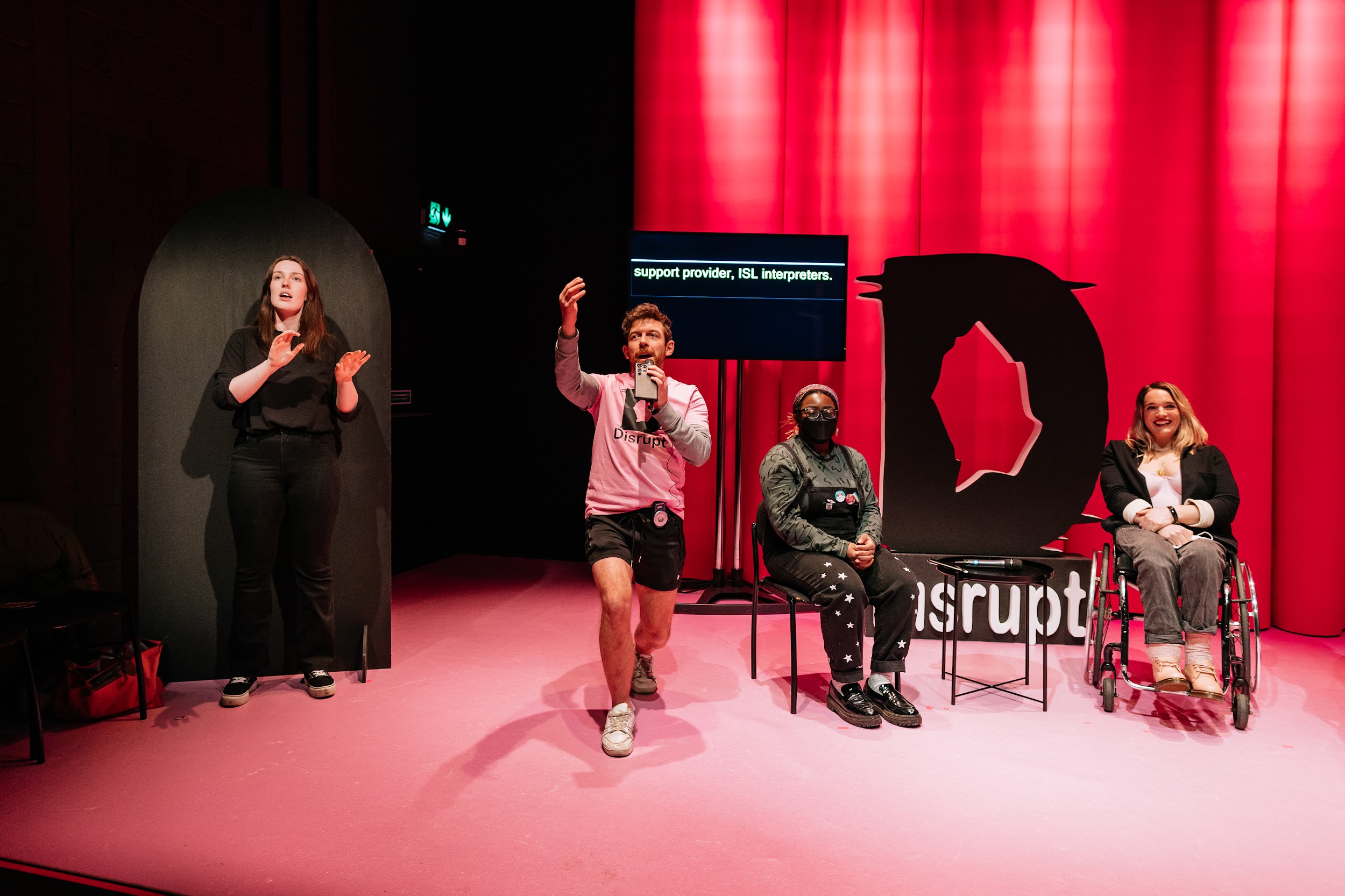 Four figures on the Disrupt festival stage. An ISL interpreter, spotlit. Festival organiser Alan James burns, who is standing gestures with animation toward the audience. Seated is Blezzing Dada, wearing Black dungarees and a facemask. Next sits writer Louise Bruton who is beaming radiantly. The scene is lit in hues of pink. Photo by Simon Lazewski.