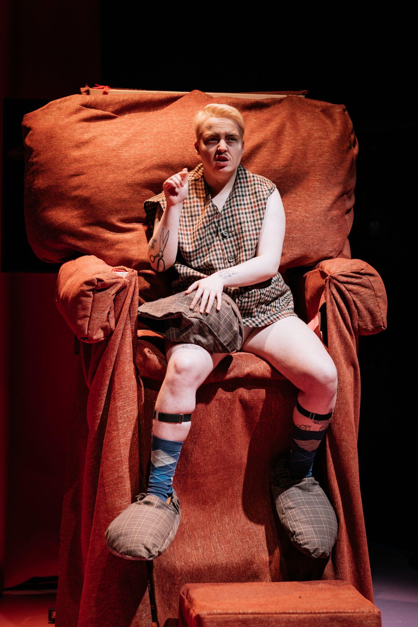 Them Fatale, a character, sits on a comically oversized chair in a deep orange, facing the camera. They are a person with short fair hair and light skin. They are wearing a brown, orange, and cream tartan plaid baggy waistcoat, with black straps around their lower legs and oversized slippers in similar colours and material. They are holding an object made of the same materials on their right leg. Photo by Simon Lazewski.