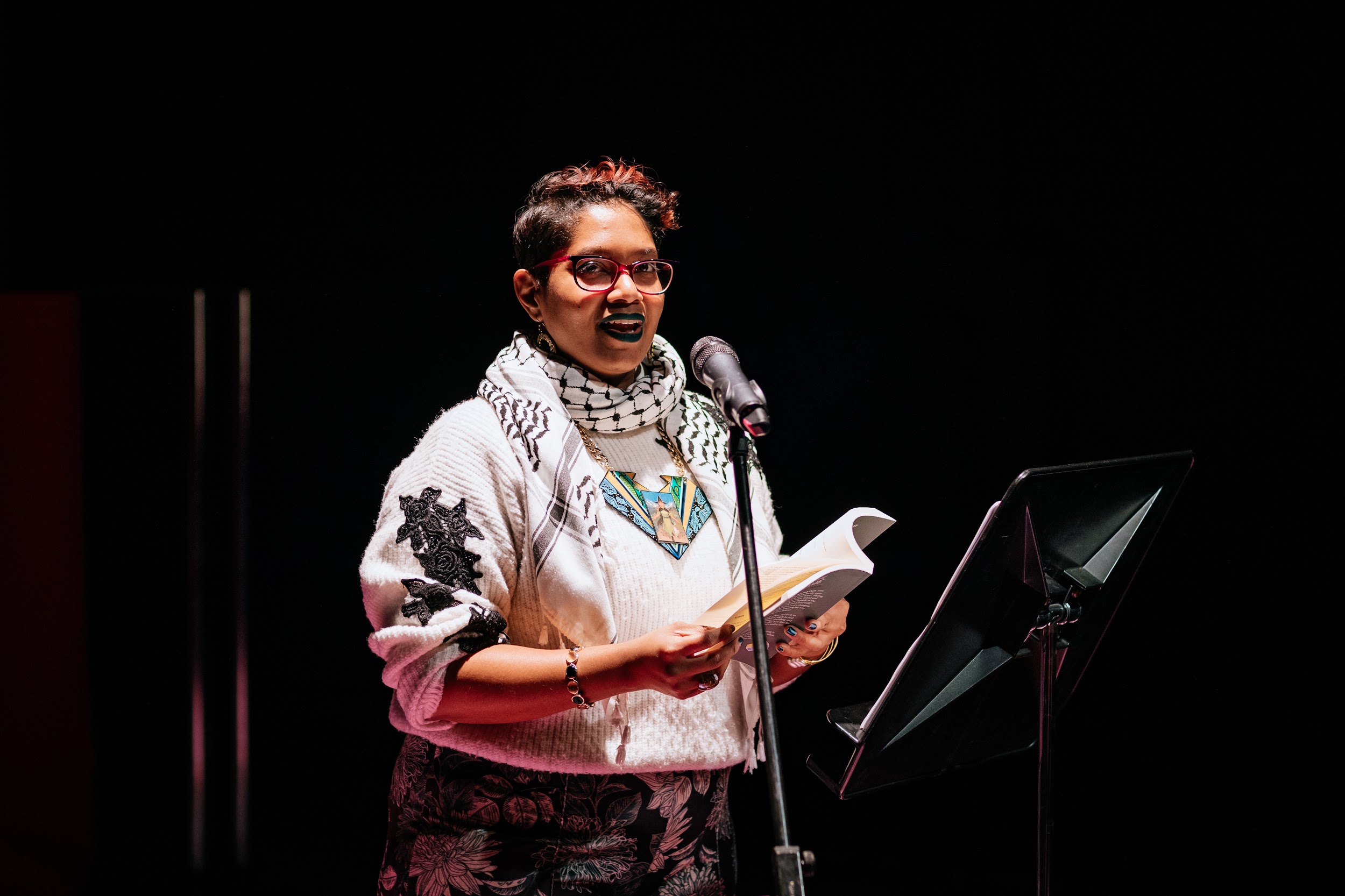 Poet Chandrika Narayanan- Mohan stands spotlit on a stage. She is reading from a book, her gaze meeting us. She has a smile on her face. She is dressed in a white jumper and wears a Palestinian keffiyeh scarf. Photo by Simon Lazewski.