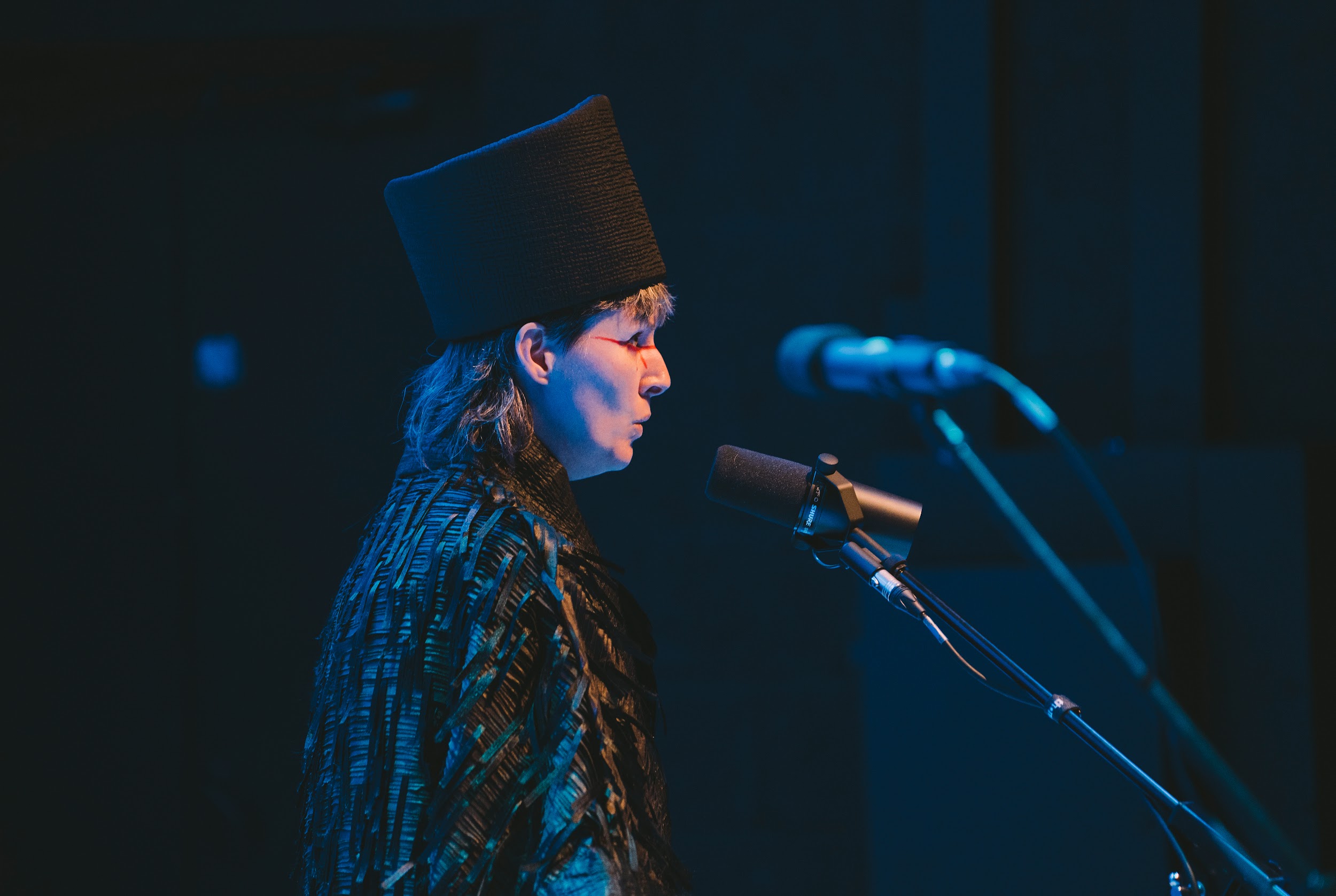 Performance artist Suzanne Walsh stands on a stage. They are dressed in a cloak of black ruffles and a tall head-dress. Graphic red makeup around their eyes. They hold their mouth in a puckered formation, as if whistling. They are lit in tones of blue. Photo by Leanne Sullivan.