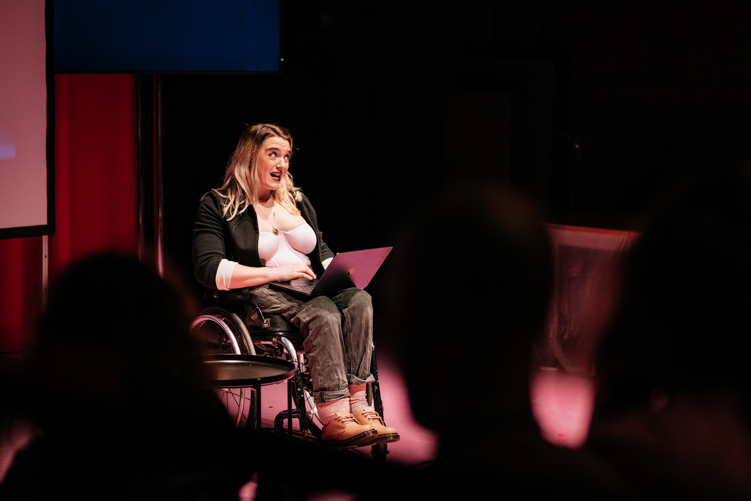 An image of artist Louise Bruton performing. Louise is sitting in a wheelchair in the centre of the photo on stage, she has a laptop on her lap and is looking out to the audience. She is a white woman with long blonde hair and wears a pink top and a black blazer. The foreground of the photo shows the dark silhouette of the audience that frames the image. The stage is lit in pink and red hues. Photo by Simon Lazewski.