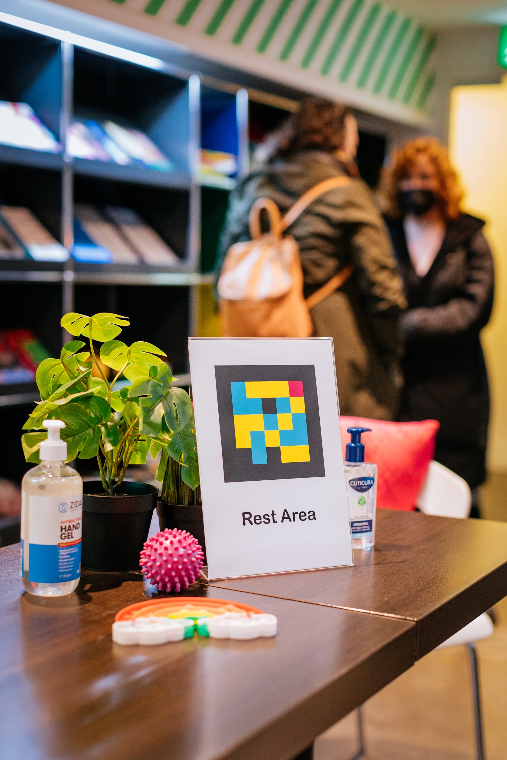 In the lobby of Project Art Center is a sign which is propped up on a table. The sign reads “Rest Area” it also has a Navicode image on it. Other objects on the table include. A plant, two bottles of hand sanitiser and two stim toys, one the shape of a rainbow, the other a pink ball.