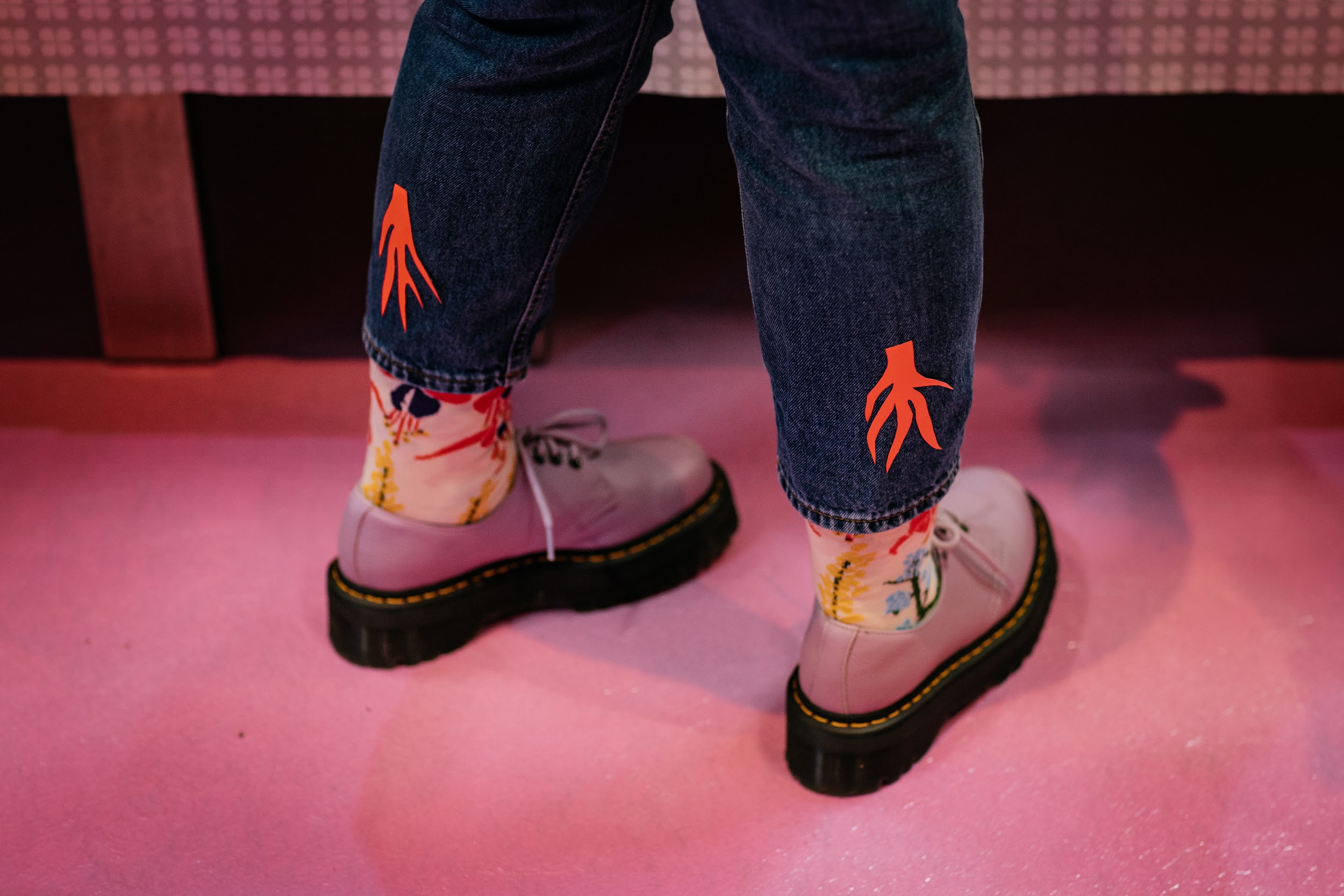 A person captured from the knees down. They stand on a pink carpet. They wear blue jeans, floral socks and pink Dr Martin shoes. On the back of their calves are paper cutouts. Bright pink in colour, the shape flame or floral like. Photo by Simon Lazewski.