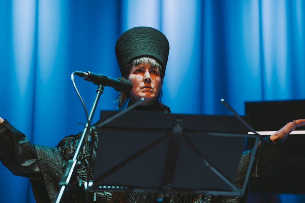 Performance artist Suzanne Walshe in action upon a stage. They are standing in blue lighting. They wear ceremonial dress, a ruffled black cloak, birdlike, with a tall headdress. Suzanne stands with a comically serious look on their face. Suzanne’s arms are spread, as if they are about to take flight. Photo by Leanne Sullivan.