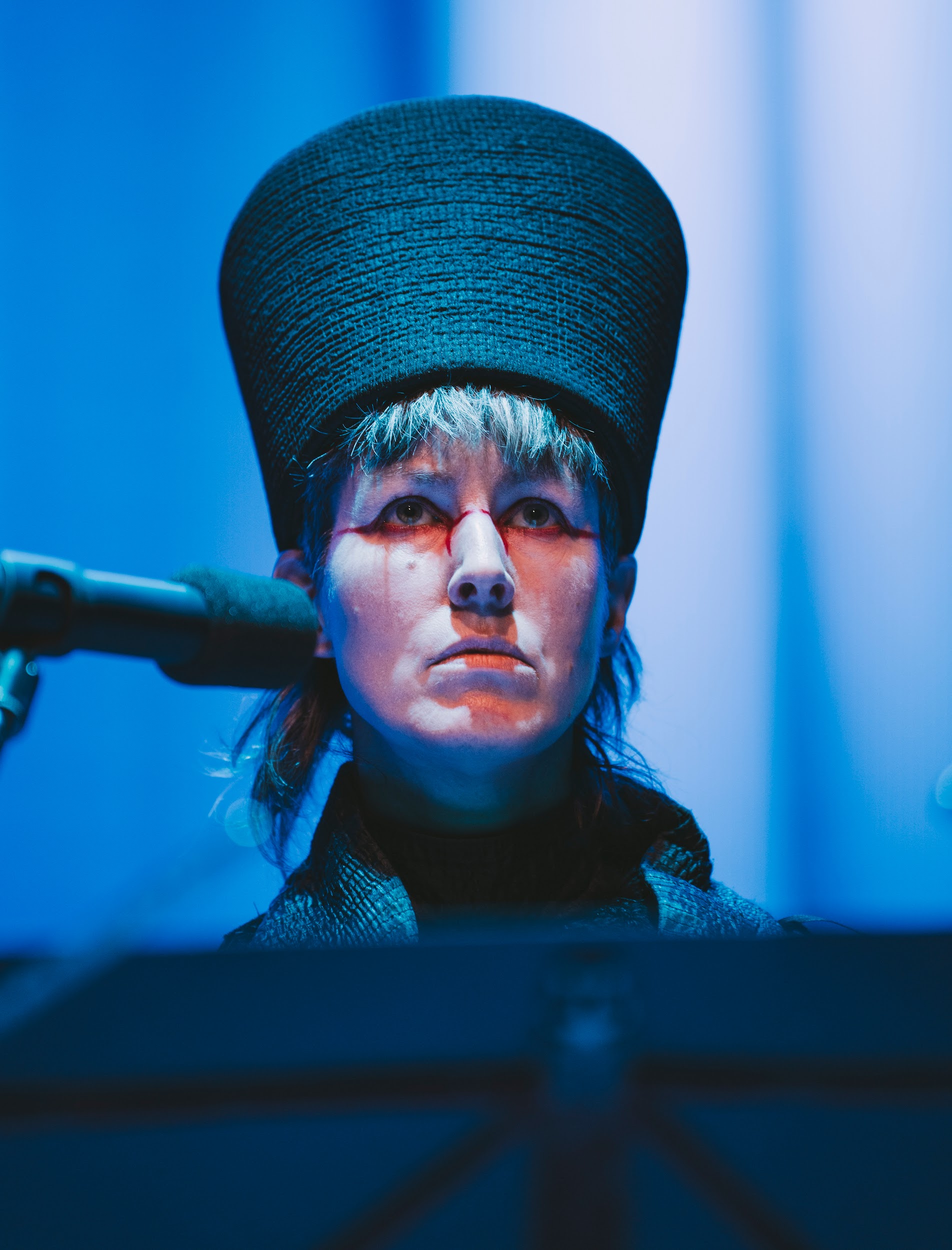 Performance artist Suzanne Walsh is shown from the head and shoulders. They wear a tall black head-dress and a steely look of defiance on their face. They are lit in tones of blue and wear dramatic red makeup around the eyes. They are lit in tones of blue. Photo by Leanne Sullivan.