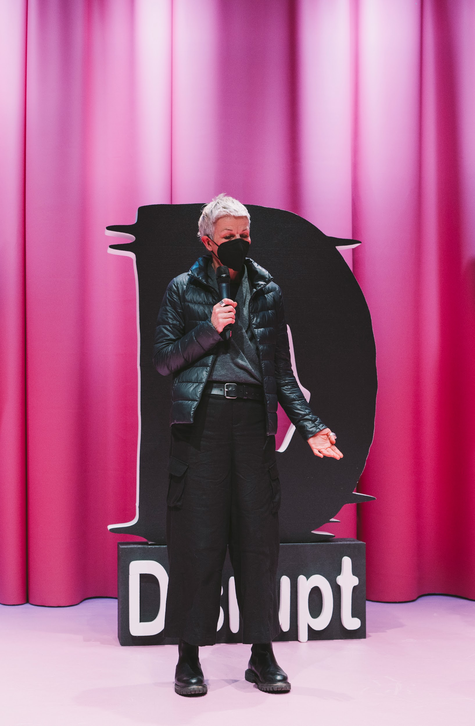 In front of a pink stage curtain, and a large styrofoam D, stands visual artist Katerine Sanky. Sanky is tall and has short grey hair. She is dressed all in black and wears a black facemask. She holds a microphone and gesticulates while speaking. Photo by Leanne Sullivan.