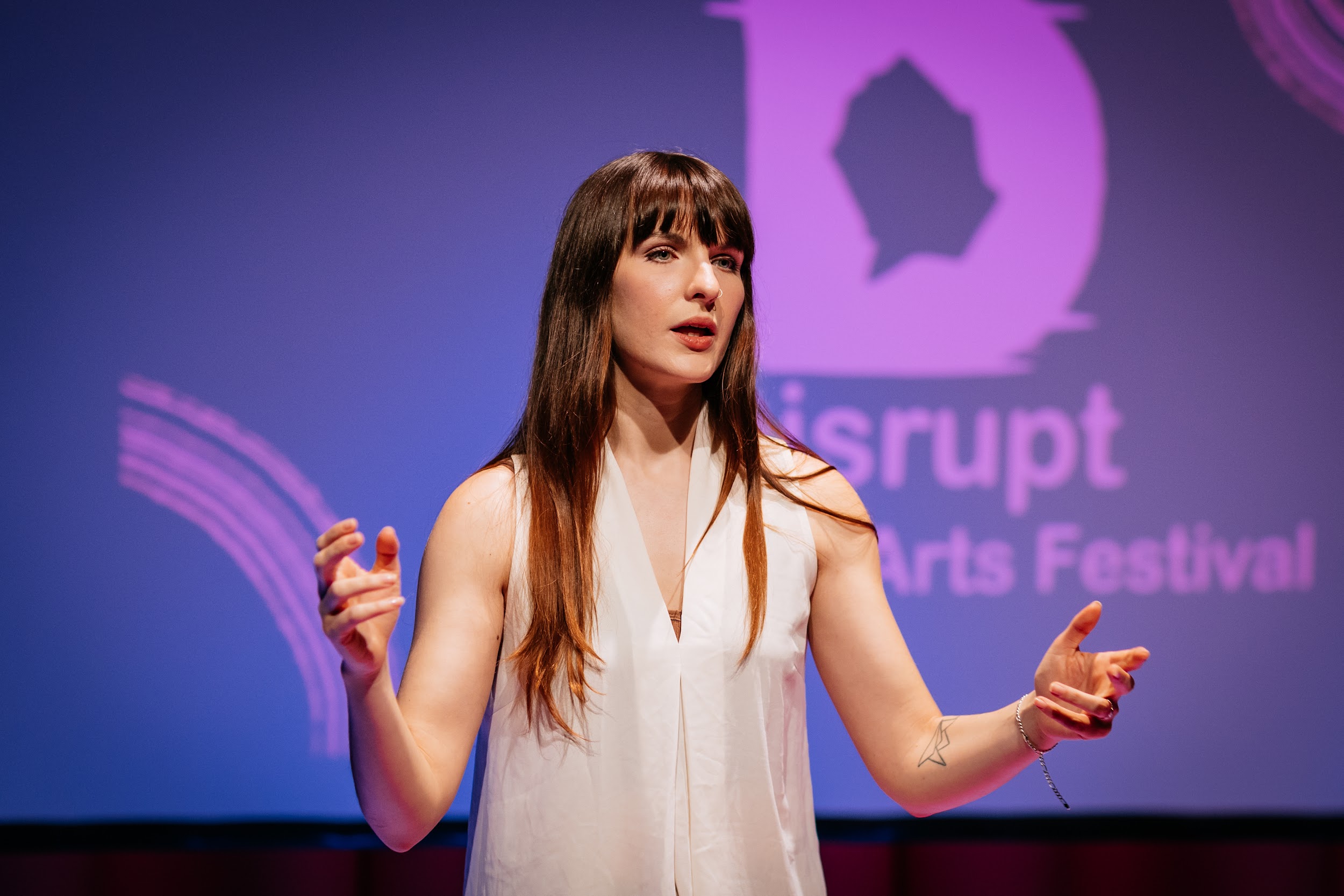 An image of artist Ali Clarke performing on stage. She stands in front of a purple background. Alison gestures outward with both hands, a searching look upon her face. Photo by Simon Lazewski.