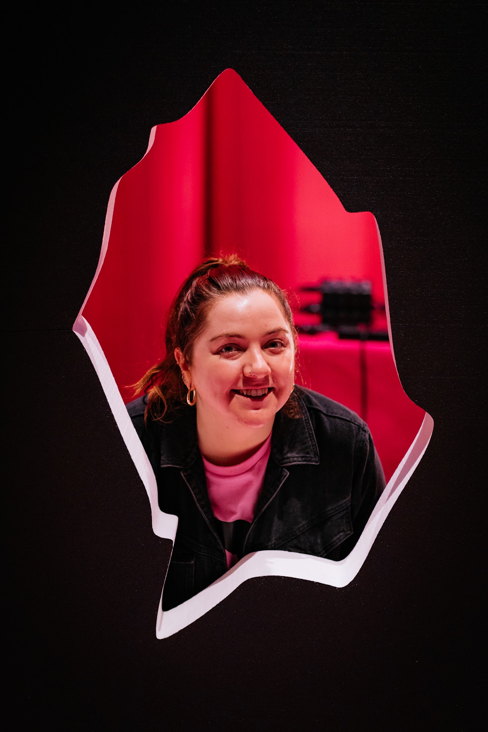 Festival crew member Laura O’ Keeffe poses for a head and shoulders portrait. The image is taken through the central cut out of the styrofoam Disrupt branding. This makes it appear that Laura is framed by a shard of broken glass. Laura is smiling to the camera.