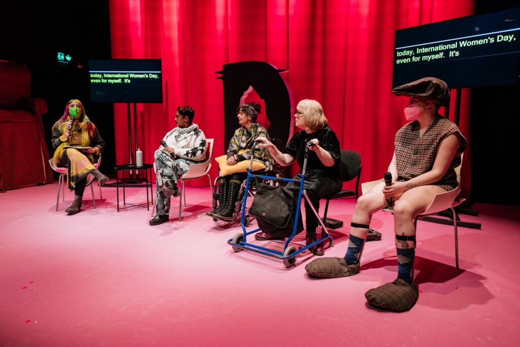 A panel discussion On a stage, lit in hues of pink. Tara Carrol hosts the conversation. They are joined by Chandrika Narayanan-Mohan, Kathy O'Leary, Mairéad Folan, and Them Fatale. Tara holds a mic. Them Fatale is dressed as a caricature of an old man. Photo by Simon Lazewski.