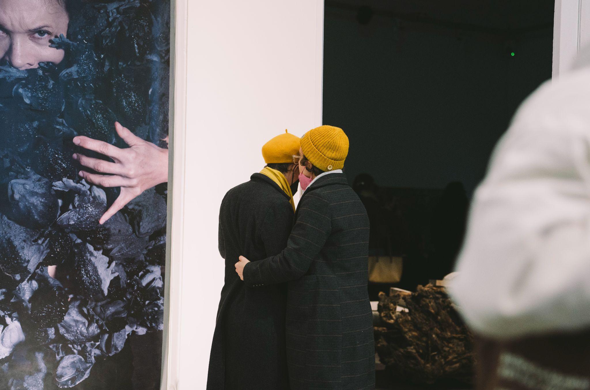 Taken from a slight distance, the image reveals two people dressed in long dark jackets and mustard-coloured hats, facing away from the camera. The person on the right wears a pink mask and has their arm around the other person. To the left of the pair is a large-scale photograph of a person with pale skin, nearly obscured by dark-coloured frogs, with only part of their face and one hand visible, mounted against a white wall. To their right is a large black screen. Photo by Leanne Sullivan.