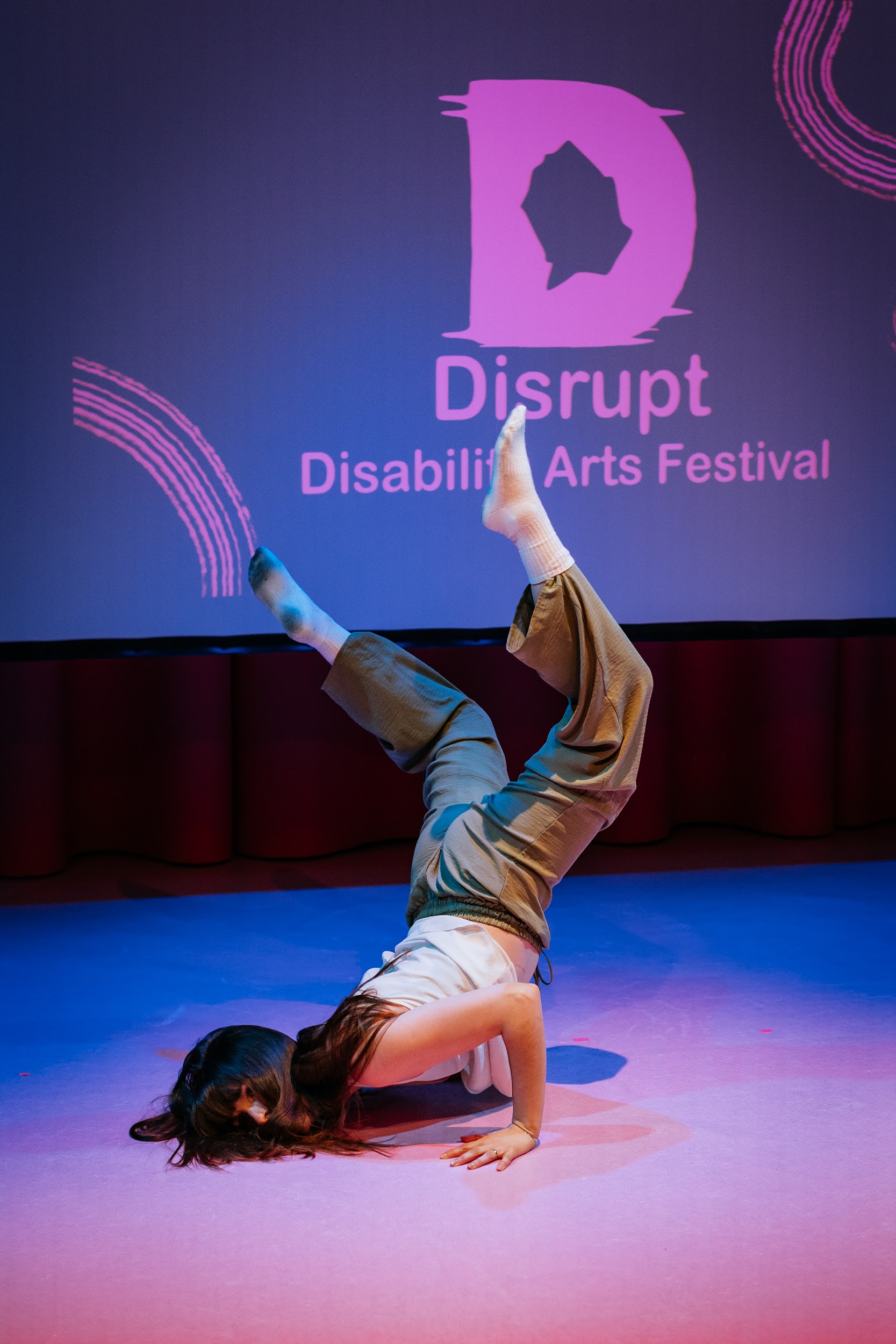 An image of dance artist Ali Clarke performing on stage. She performs a complicated move. A leap, downward, where her chest connects with the ground, legs in the air. It appears as if she will follow through with the motion to roll upward again. The scene is lit in hues of pink and blue. Photo by Simon Lazewski.