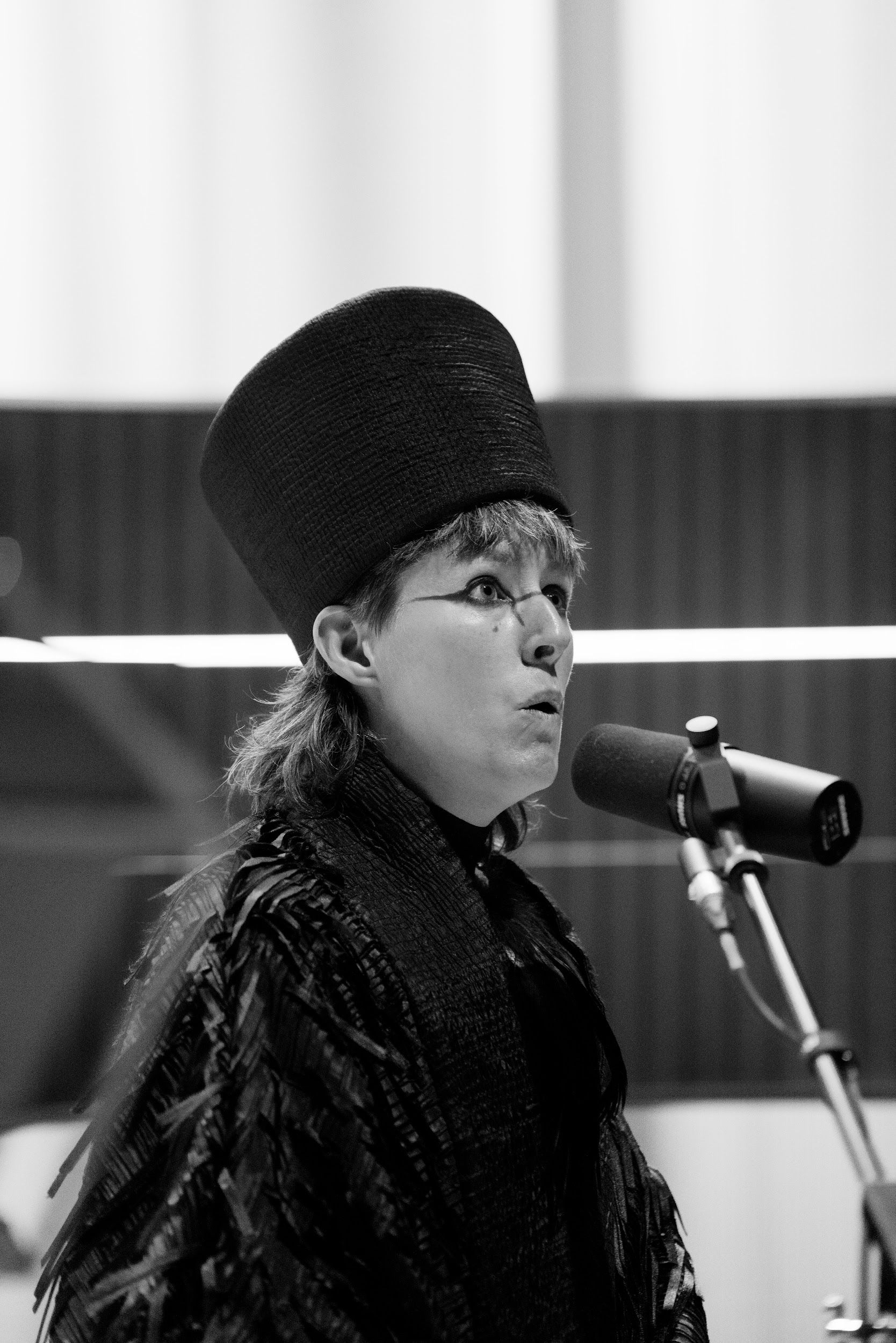 Performance artist Suzanne Walsh stands on a stage. They are dressed in a cloak of black ruffles and a tall head-dress. Graphic red makeup around their eyes. They hold their mouth in a puckered formation, as if making a hooting sound. The image is in black and white. Photo by Leanne Sullivan.