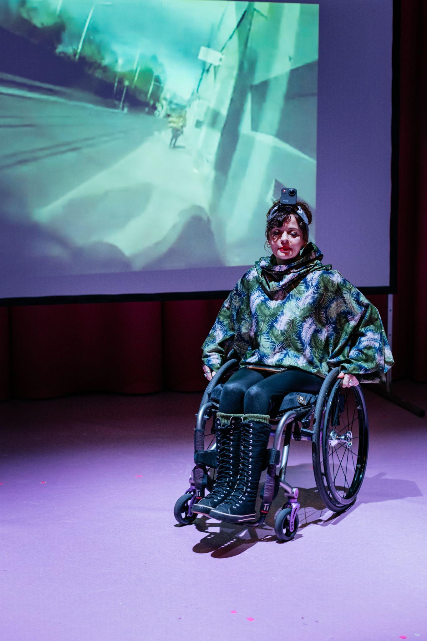 Artist Kathy O'Leary, a person with long dark hair tied with a hair bow, wearing a long-sleeved green top adorned with large green and white feathers, black shiny trousers, and long black runner boots, sits in a wheelchair positioned diagonally on a stage. They are illuminated with purple and pink hues. Behind them, a projection screen displays an image of a street scene with a person walking beside tram tracks. Photo by Simon Lazewski.