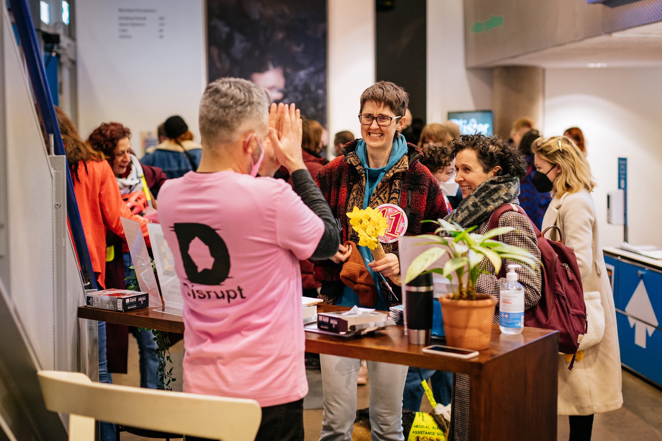 A member of the Disrupt festival crew stands behind an information point in Project Art Centre’s lobby. They are making a gesture of respect towards two feminine figures who are smiling deeply.