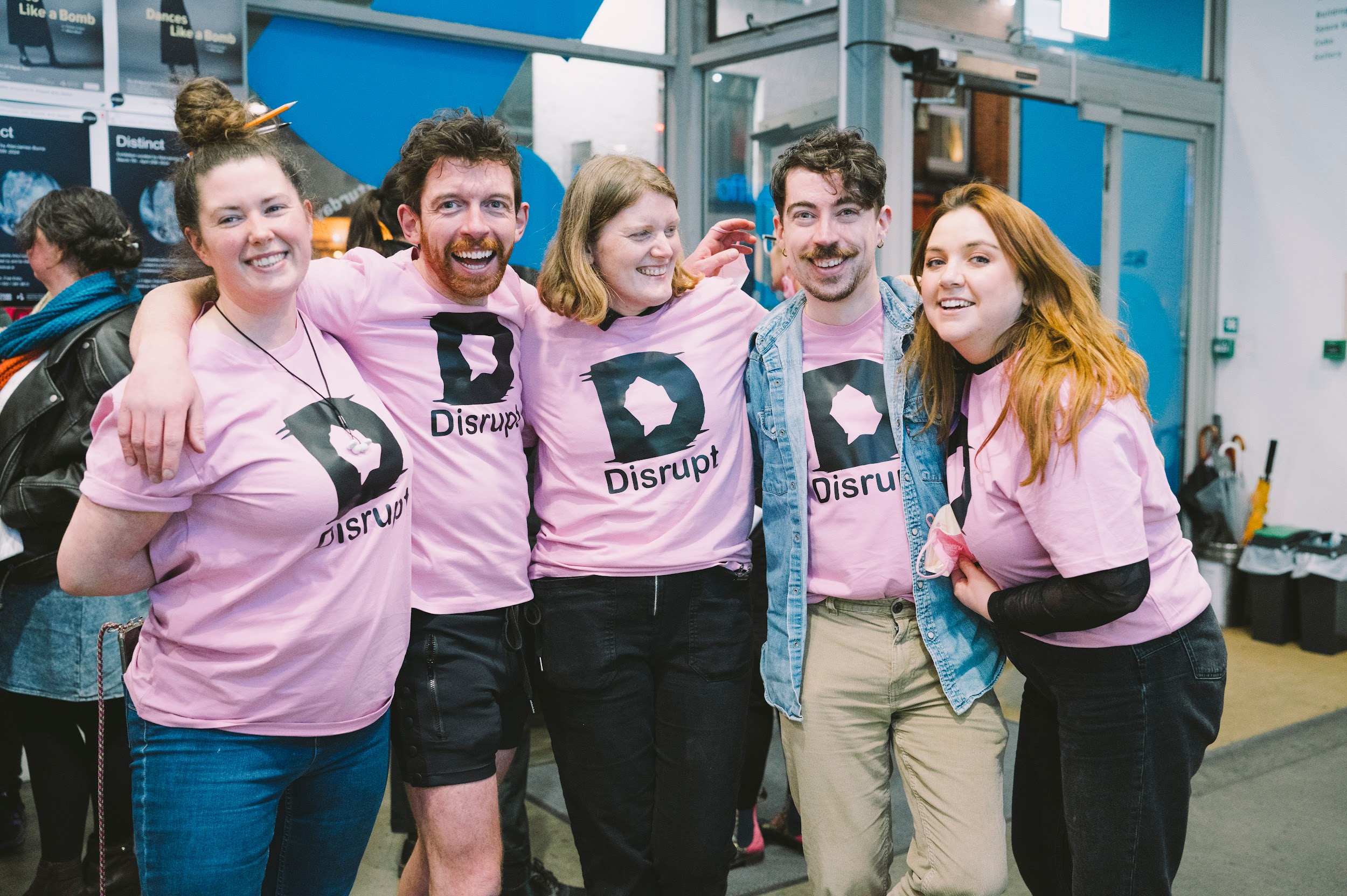 A group of people wearing pink t-shirts pose for a photograph. On each of their T-shirts is the Disrupt festival logo. The group have their arms linked around each other and all have expressions of elation. Photo by Leanne Sullivan.