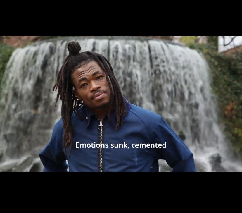 This is an image of artist Tobi Balogun’s performance. It is a still of a video art piece Tobi created for Disrupt. In the centre, we see Tobi who is a black Yoruba-Irish male. He is wearing a blue jacket with a silver zip down the front. His hair is dark in colour and in braids. The top half is styled in a bun. In the background we see a big waterfall that fills the background space. The image also includes open captions reading ‘Emotions sunk, cemented’. Photo by Simon Lazewski.