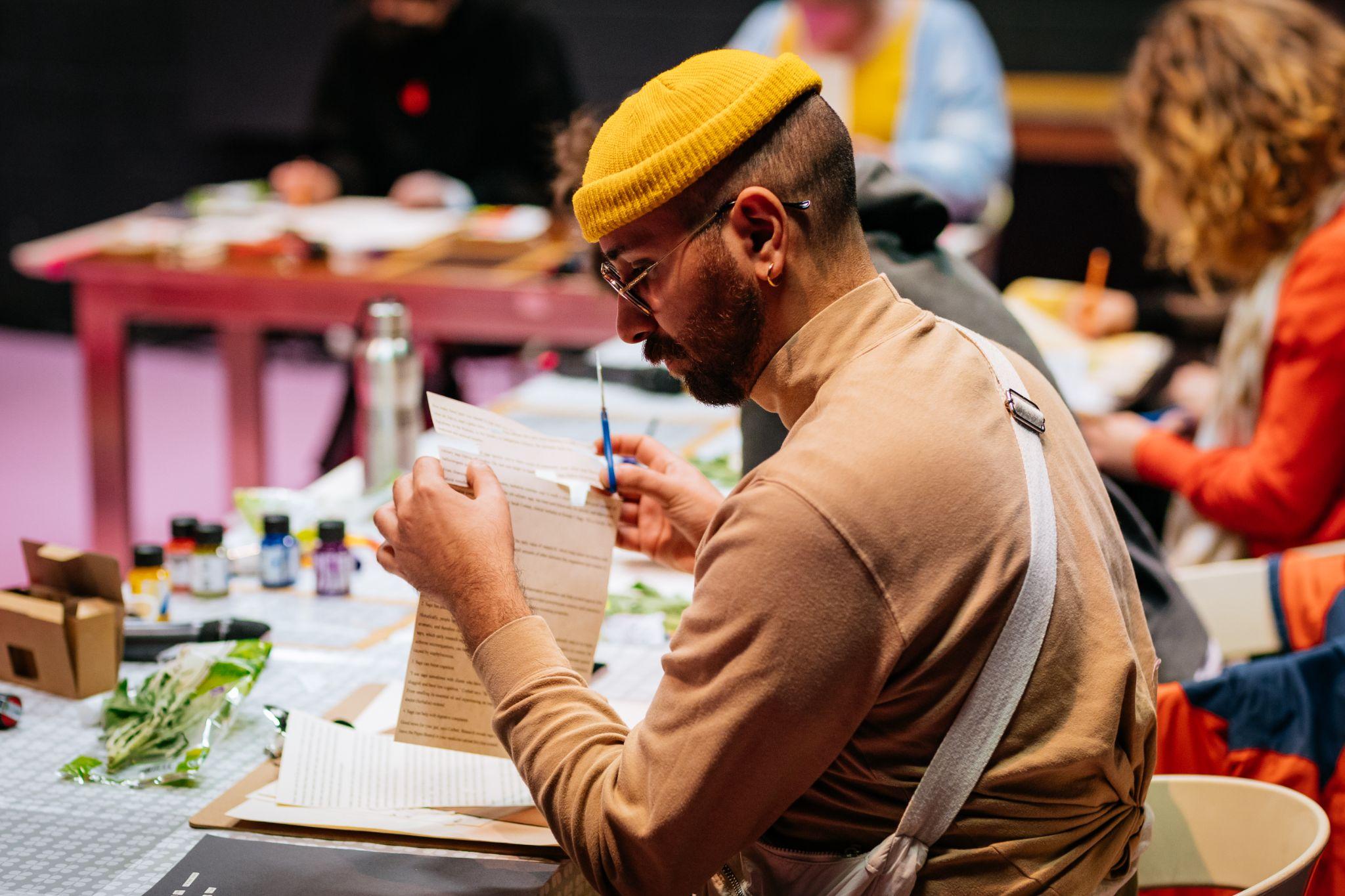 The image shows a person with short dark hair and warm skin, wearing glasses, a yellow hat, along with a light brown polo neck top. They sit at a long desk alongside other people, who are blurred in the background. The person appears to be cutting words from a page of text with scissors. On the table in front of them are various materials, including more texts, small boxes, and paints. Photo by Simon Lazewski.