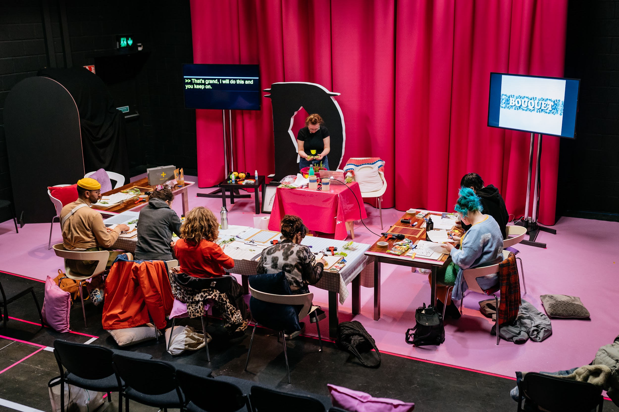 A group of six people attend a workshop by Michelle Hall. They sit at tables organised in a u formation around a centre table where Michelle stands. Michelle stands in front of a Styrofoam D, the disrupt logo. Beside Michelle is a large screen, which displays her words in real-time. On all of the tables are various art materials. The scene is captured from a downward angle. The tones are all in pink. Photo by Simon Lazewski.