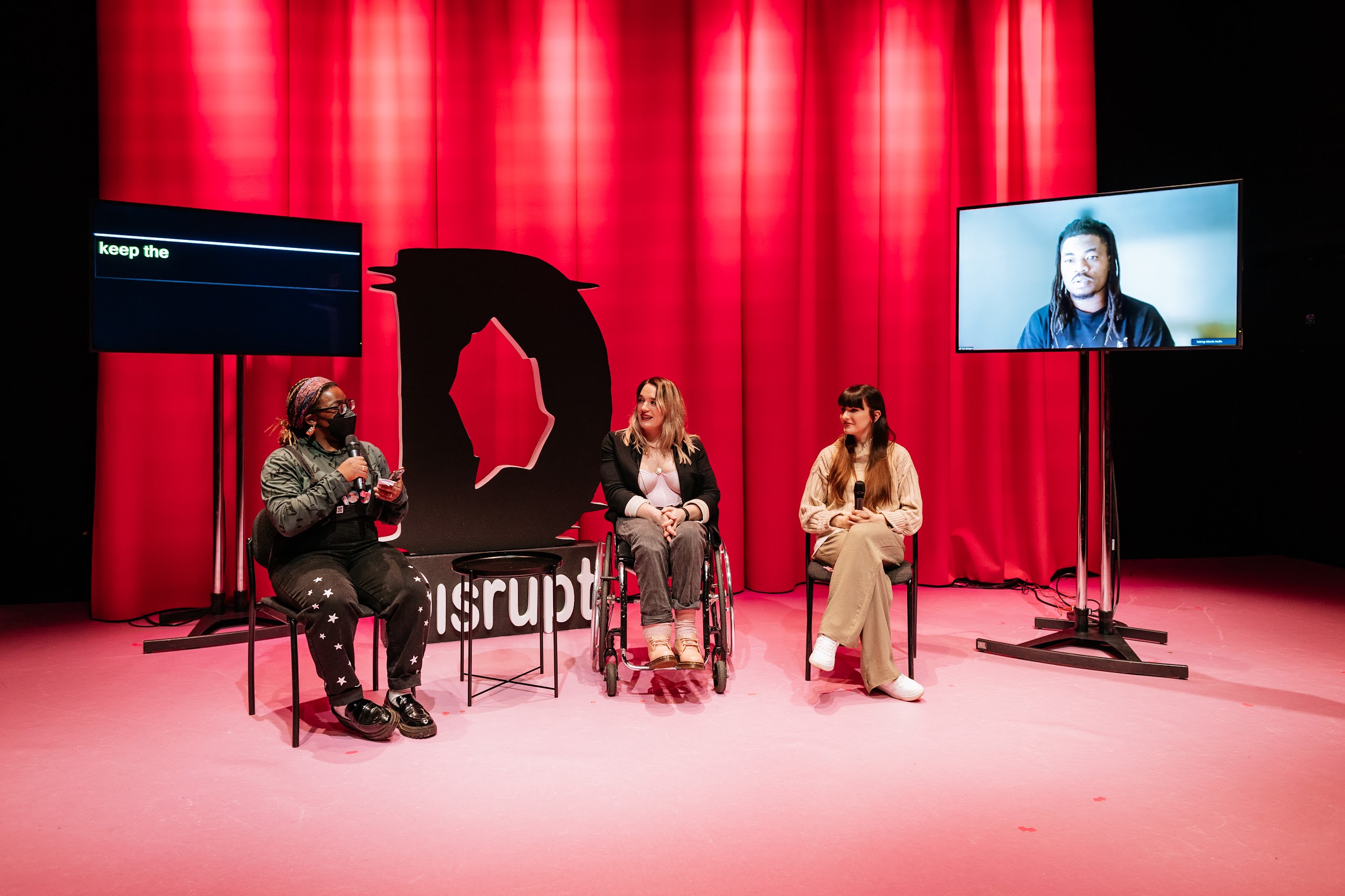 This image shows the lineup of artists for the panel discussion. The stage is well lit in the background and we see a dark pink curtain backdrop. To the left there is a monitor high on a stand being used for captions and to the right on the image we see another monitor high on a stand showing a video linked Zoom caller. This is artist Tobi Balogun and he is Yoruba-Irish with black skin and shoulder length dark hair. In the centre of the image we have 3 individuals sitting in chairs facing the audience. Far left we have Blezzing Dada, a person with dark skin who is wearing a green long sleeve t-shirt and black dungarees. Next we have Louise Bruton who is sitting in a wheelchair. She is a light skinned woman with long blonde hair and wears a pink top and a black blazer. Next we have Ali Clarke who is a light skinned woman with long brown hair and a fringe. She wears cream trousers and a cream jumper. In the background left of the photo we see a large black foam cutout of the Disrupt logo, which is a D. Photo by Simon Lazewski.