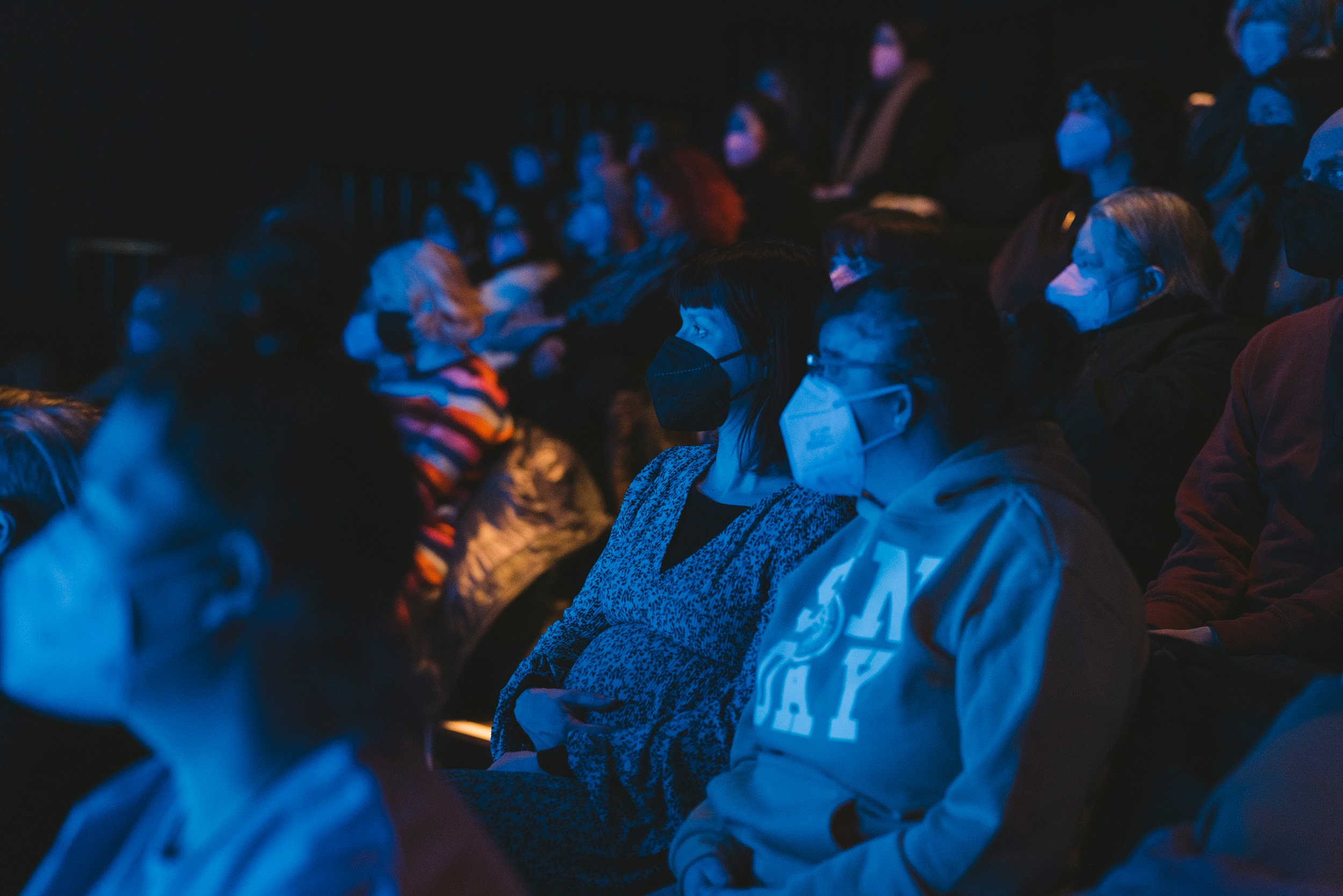 Artist Vyonne Condon sits amongst a crowd looking towards a stage. They wear a facemask and hoodie. They are lit in hues of blue. Photo by Leanne Sullivan.