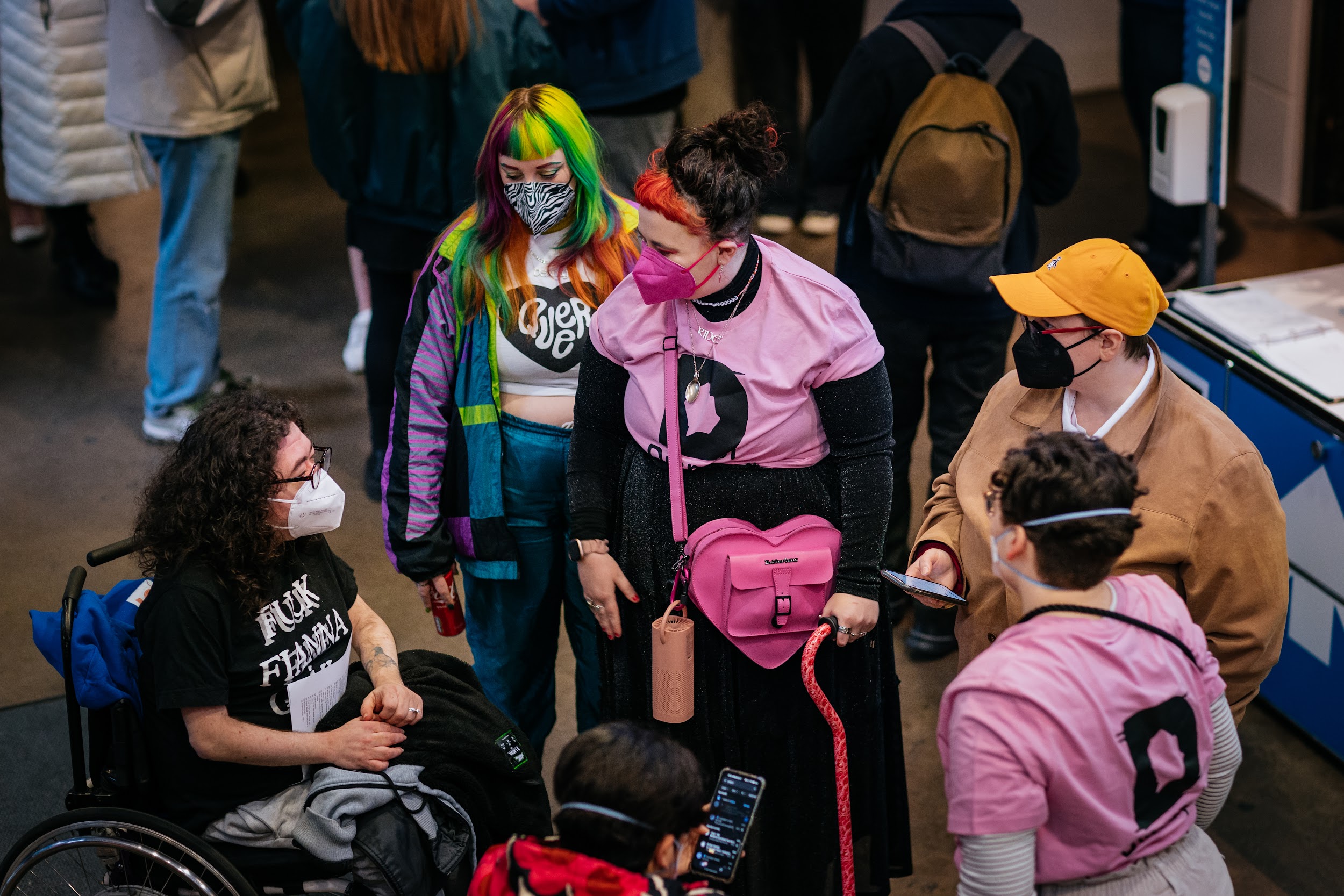 A group gather in the lobby of project arts centre. All of them wear facemasks. Two of the people wear pink disrupt t-shirts. One of the group is using a cane, another uses a wheelchair. The image is taken from above and is looking down on the scene.