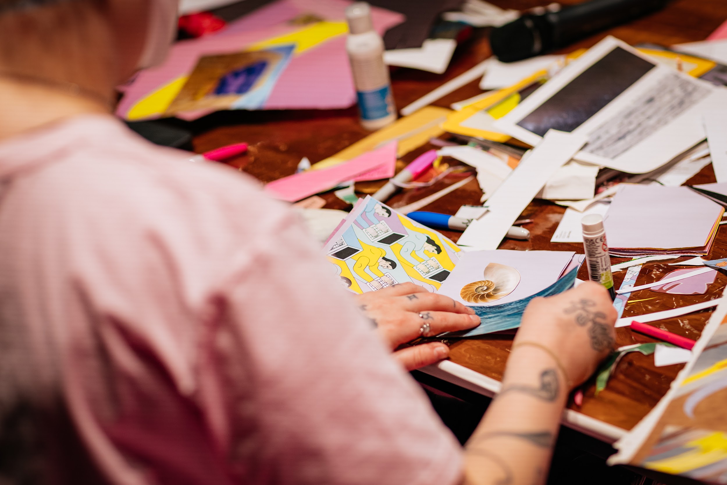 A figure wearing pink, is sitting at a desk which is covered in coloured paper and magazine cuttings. They are making a zine, and are putting a collage of a shell and an image of the sea together. Photo by Simon Lazewski.