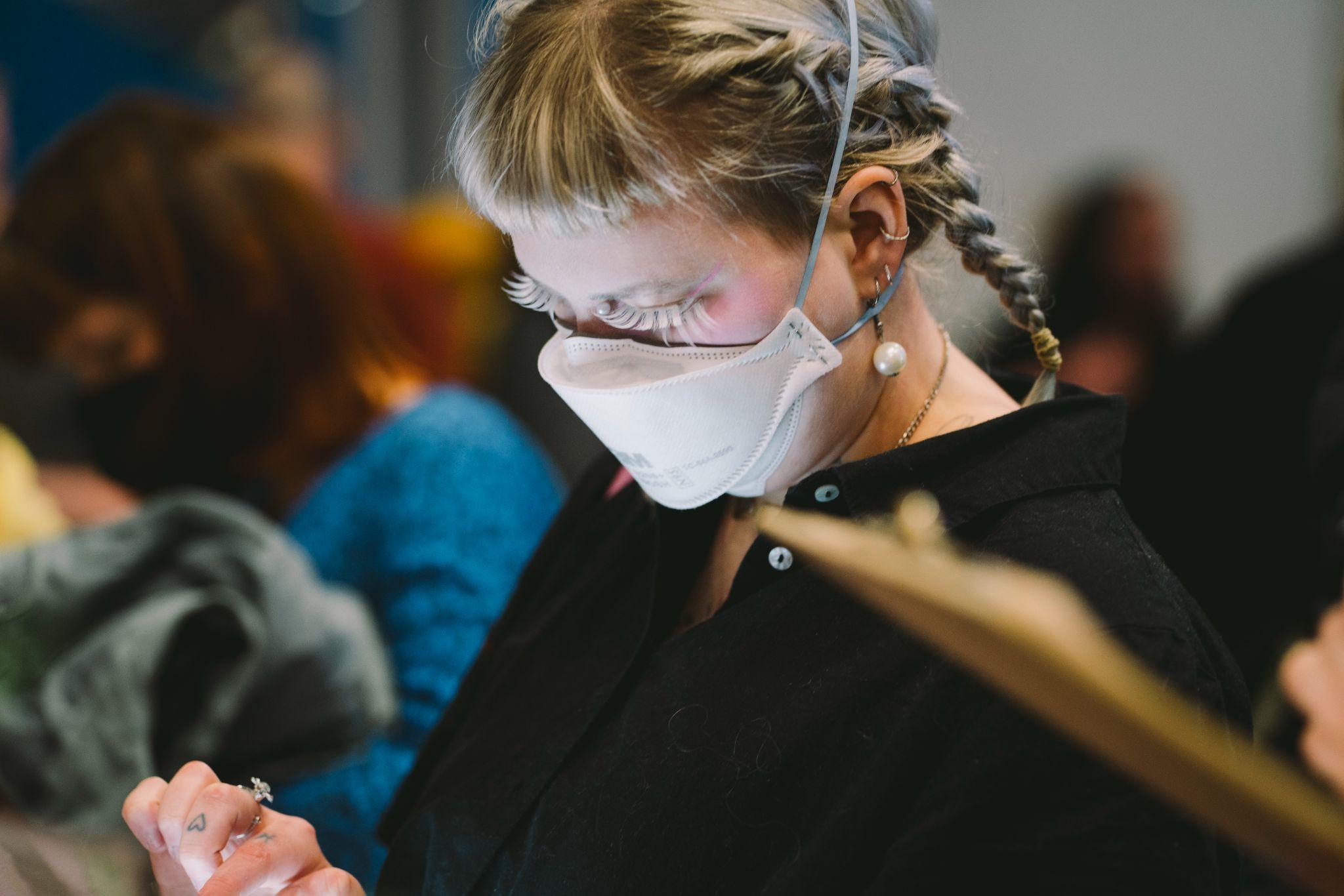 A person with fair hair styled in a plait and fringe, wearing glasses and a white mask. They are positioned in the foreground, in focus, and looking down towards a screen while tapping it with their hand. Although the screen is out of the frame, its light shines onto their face. Additionally, an out-of-focus brown object resembling a clipboard is visible in the foreground. Behind them, a group of people is out of focus. Photo by Leanne Sullivan.