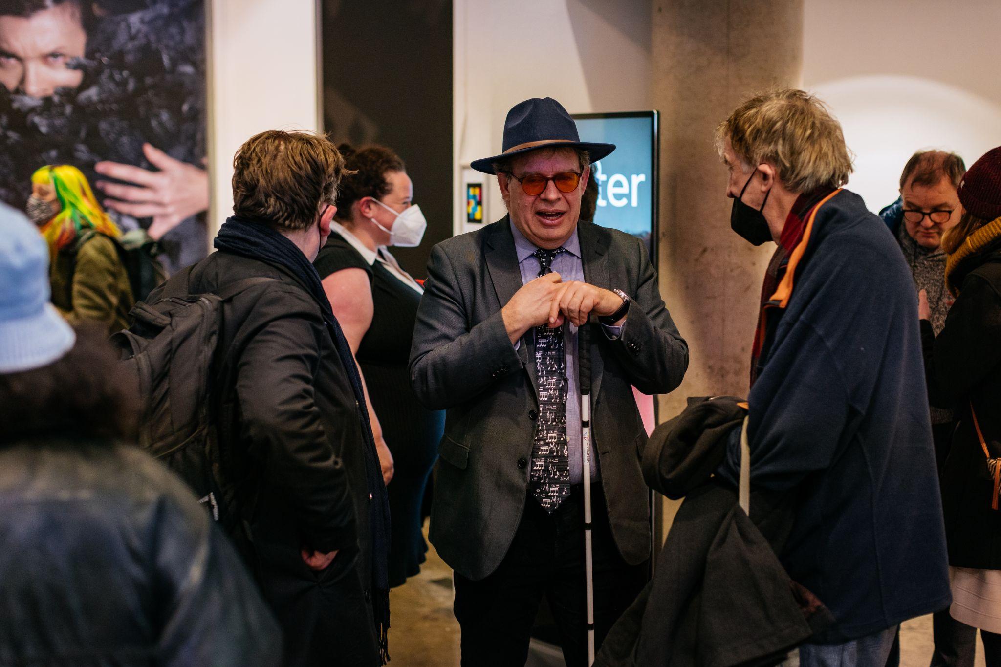 A group of people socialise in an exhibition space, with three individuals at the centre of the image engaged in discussion. The person most visible in the image faces forward, resting both hands on a cane, while two others surround them. Various artworks are mounted on the walls in the background. Photo by Simon Lazewski.