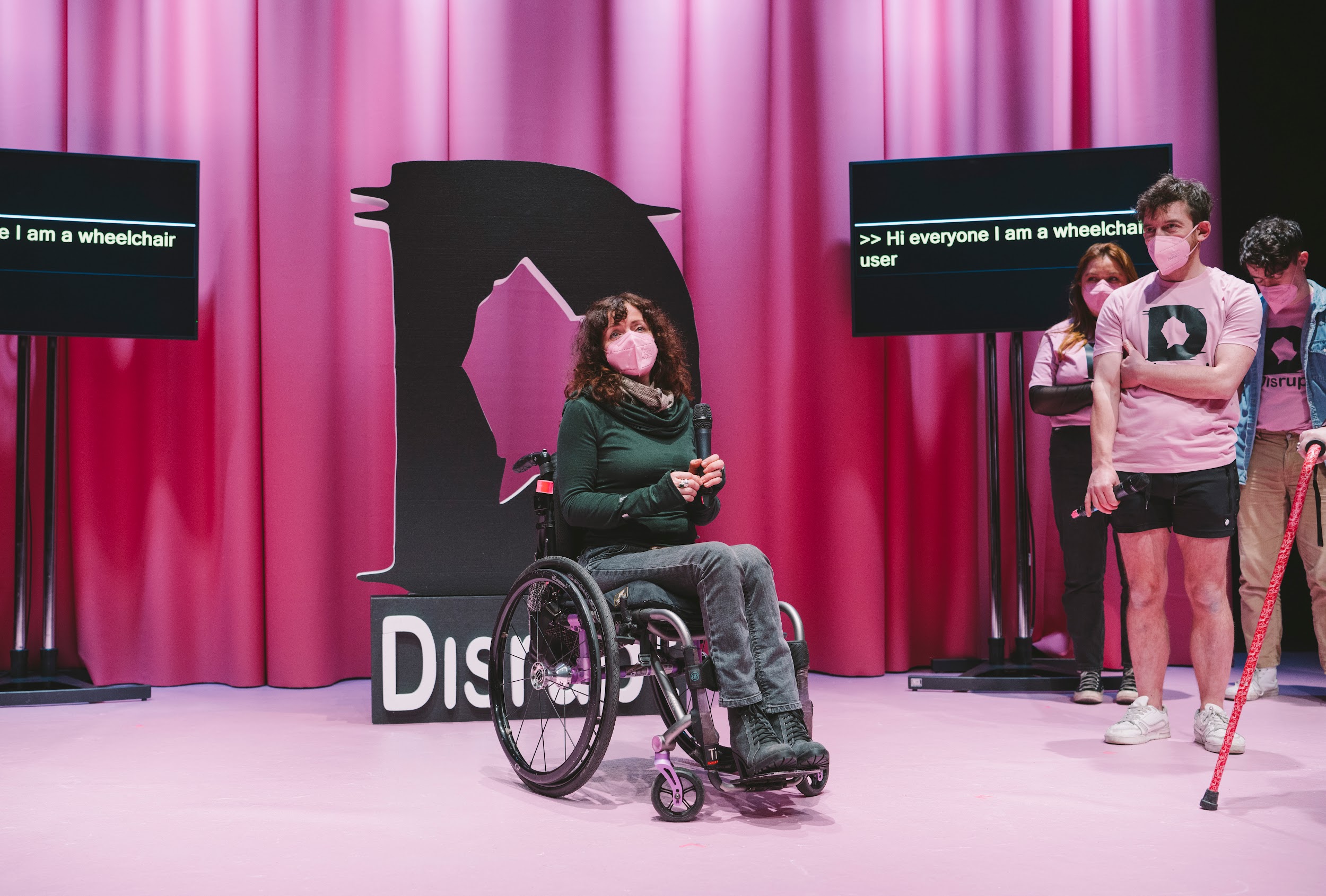 Artist Kathy O’Leary on the Disrupt festival stage. Kathy wears a green jumper, black jeans, black boots and a pink face mask. Kathy is a wheelchair user. She holds a microphone. She gazes outward towards the audience, with a look of mesmerisation. Behind Kathy, the stage is dressed in pink. A pink curtain and carpet. There is the Disrupt logo, a large D, in styrofoam. There are two screens which show Kathy’s captions. In the wings festival crew look on encouragingly. Photo by Leanne Sullivan.