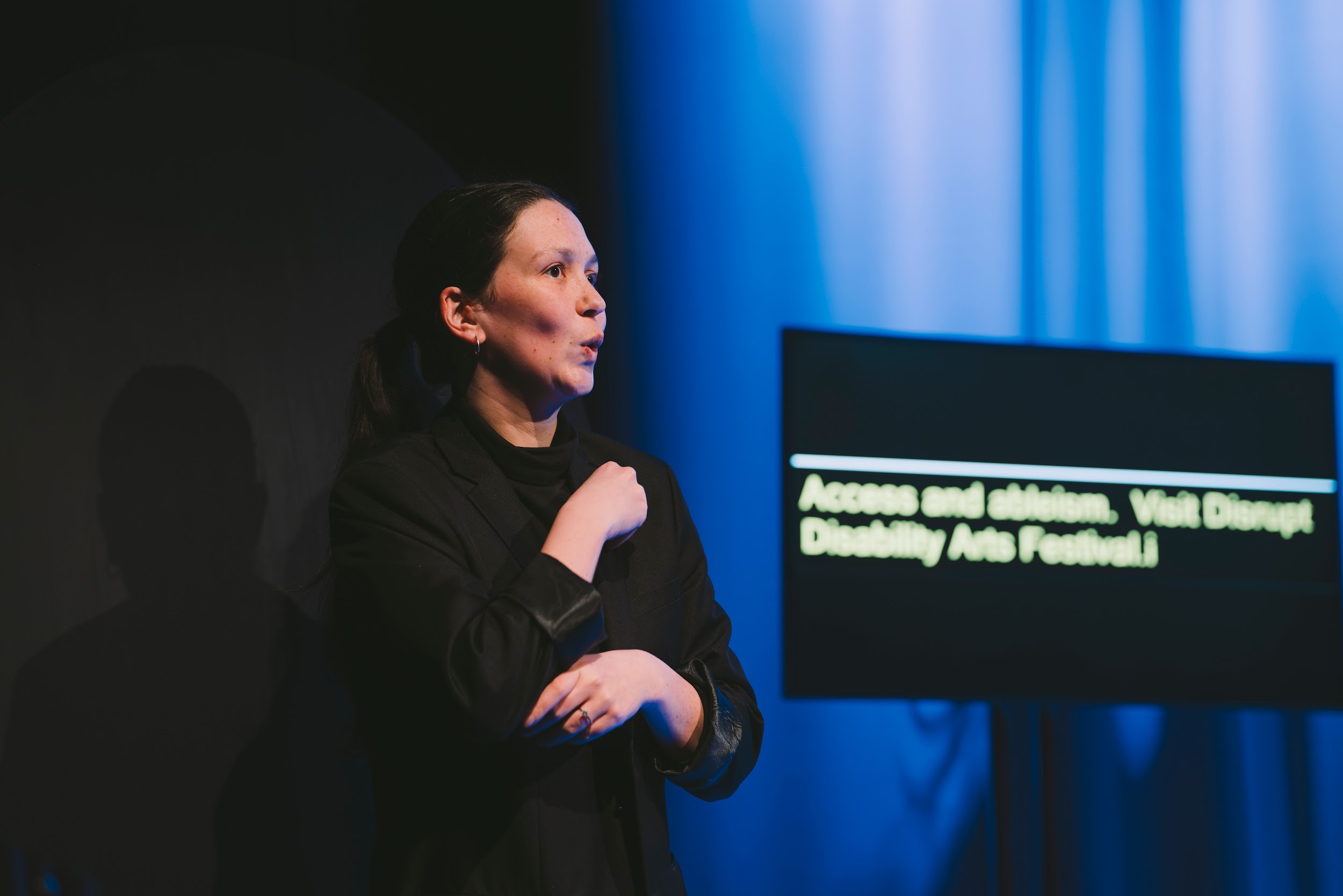 An ISL interpreter on the Disrupt festival stage. They gesture towards an audience. Behind them a screen shows captions. Photo by Leanne Sullivan.