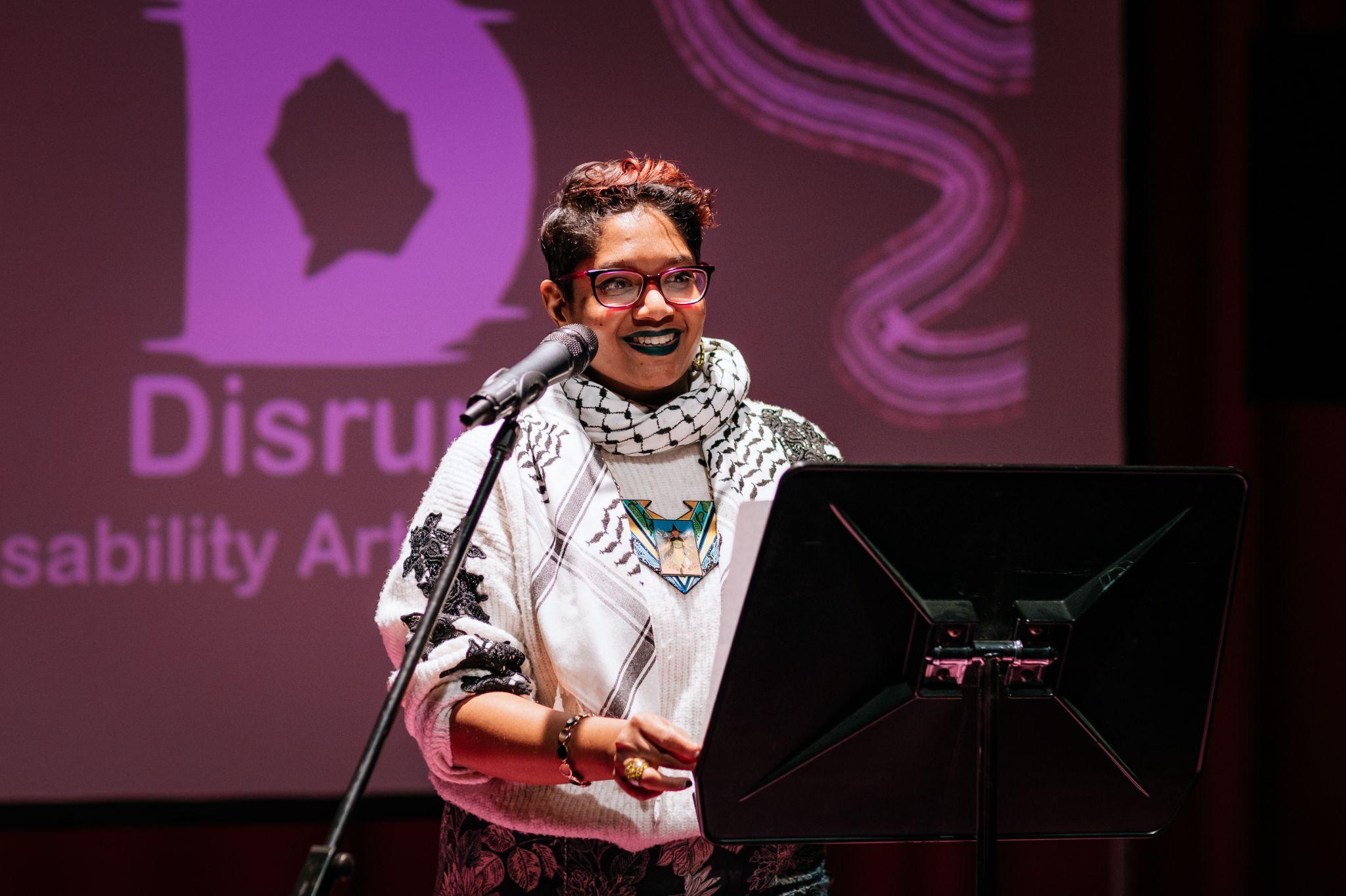 Chandrika Narayanan-Mohan, a person with short, dark hair and dark skin stands illuminated in front of a microphone, speaking while holding a book in their hands. They are wearing a light-coloured jumper and scarf featuring various patterns, and glasses. In front of them is a document holder on a stand. Behind them is a projection screen on a stand displaying the logo for Disrupt Disability Arts Festival, which is partly covered and out of the frame. Photo by Simon Lazewski.