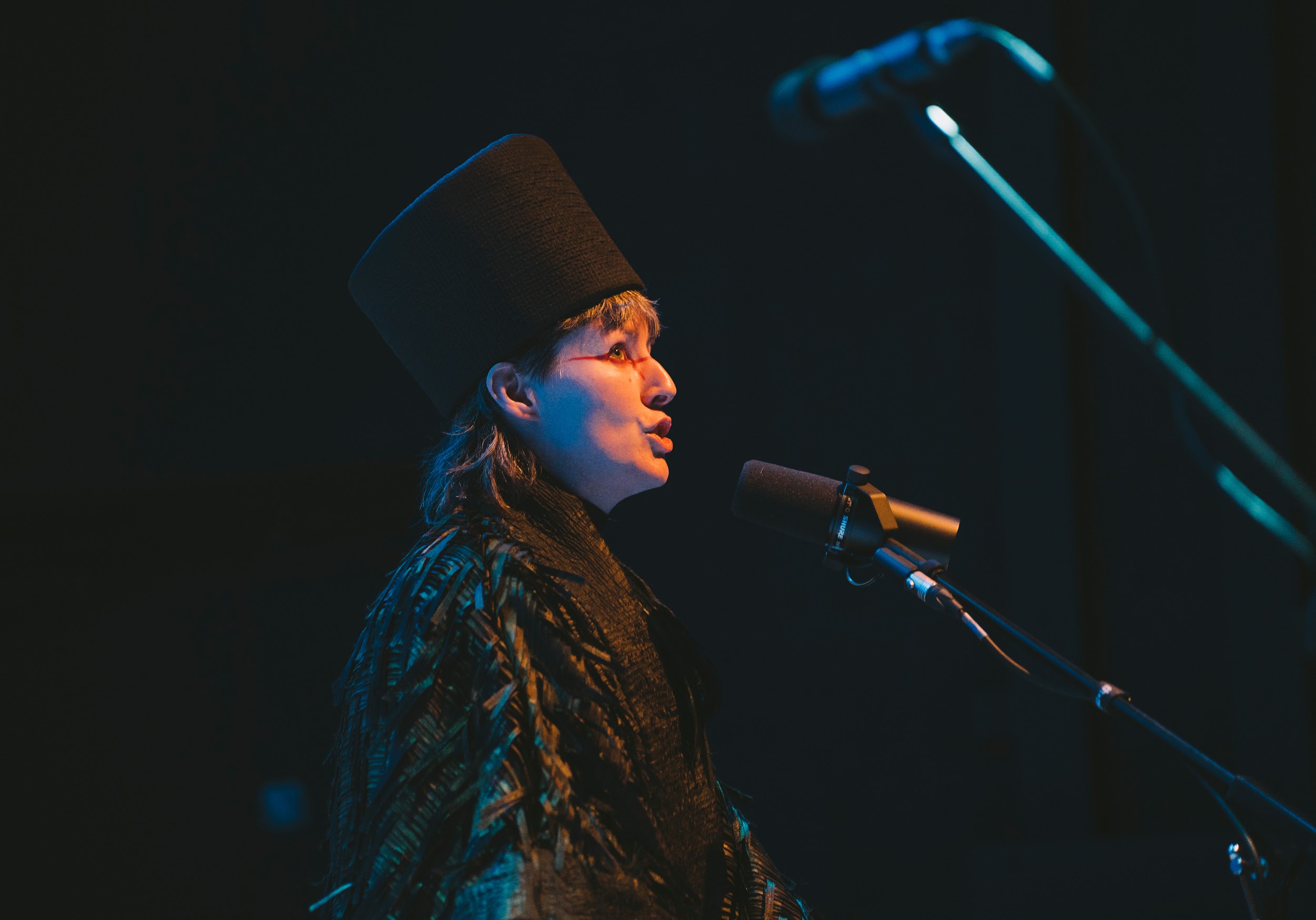 Performance artist Suzanne Walsh stands on a stage. They are dressed in a cloak of black ruffles and a tall head-dress. Graphic red makeup around their eyes. They hold their mouth in a puckered formation, as if chirping. They are lit in warm tones. Photo by Leanne Sullivan.