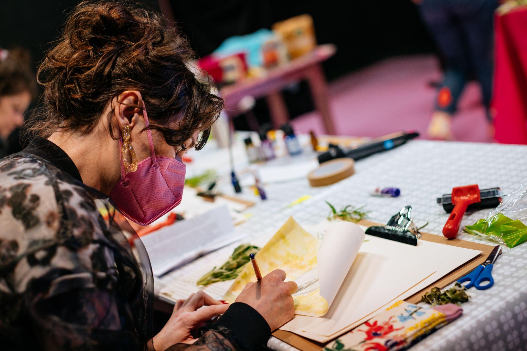 A person sitting at a desk, captured over shoulder. They are engaged in drawing. The person has long dark hair and light skin. They are wearing a dark and light patterned top along with a bright pink mask. On their table are various materials, including a clipboard with blank sheets, leaves, and scissors. In the background, other crafting materials are visible. Photo by Simon Lazewski.