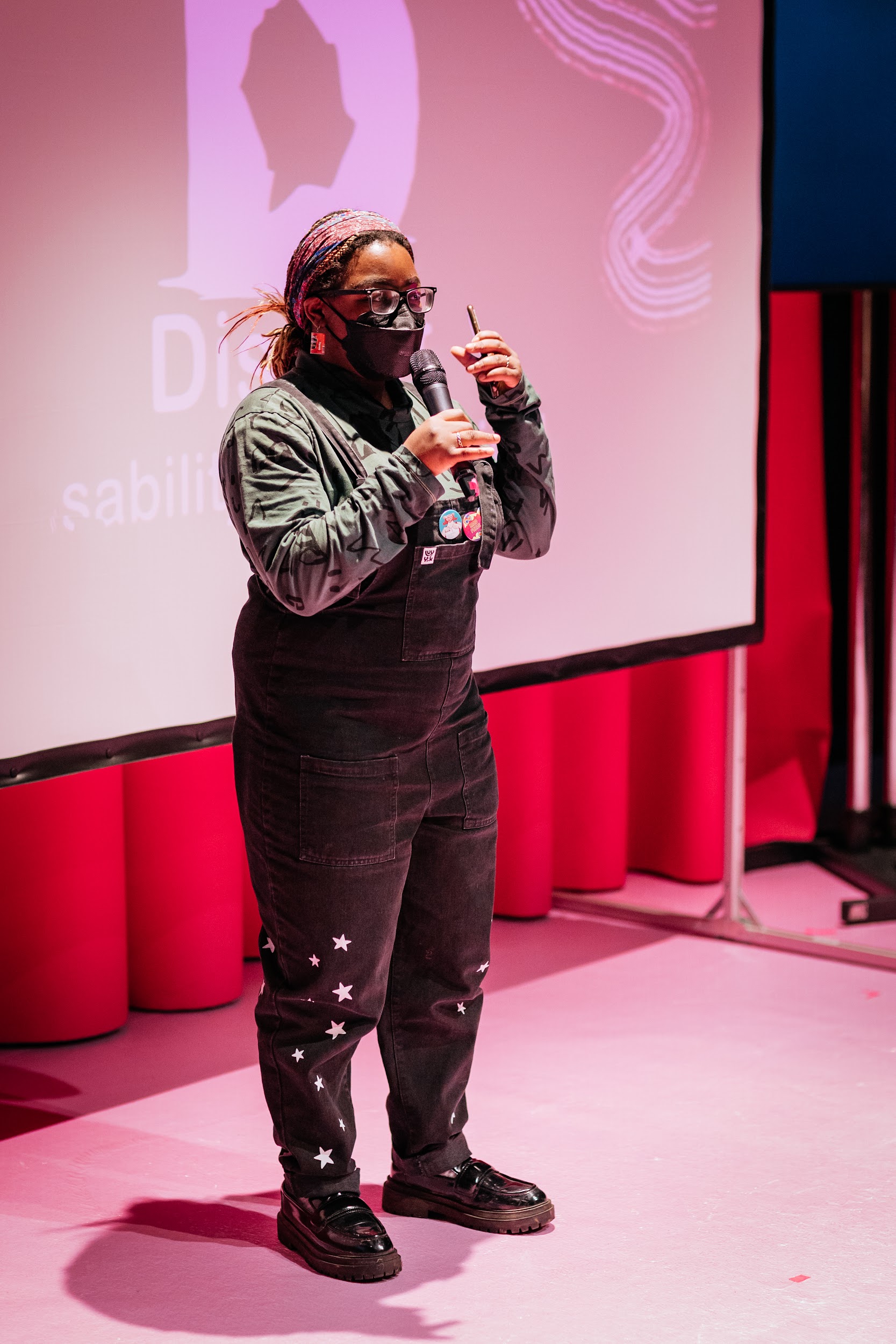 Blezzing Dada is on the stage at Disrupt Disability Arts Festival. They wear black dungarees and a black face mask. Behind Blezzing is a projection screen which has the festival branding illuminated on it.