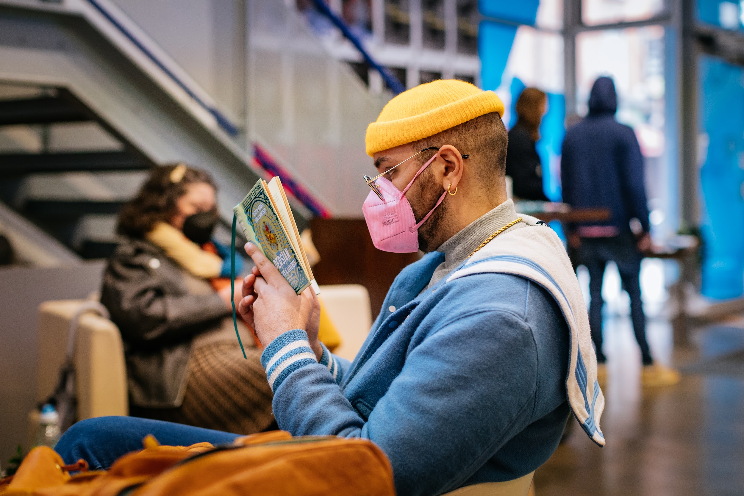 A man reads some literature in a lobby. He is wearing a blue nautical style jacket, a small yellow beanie and a pink face mask. In the background we can see a woman who is also sitting and reading.