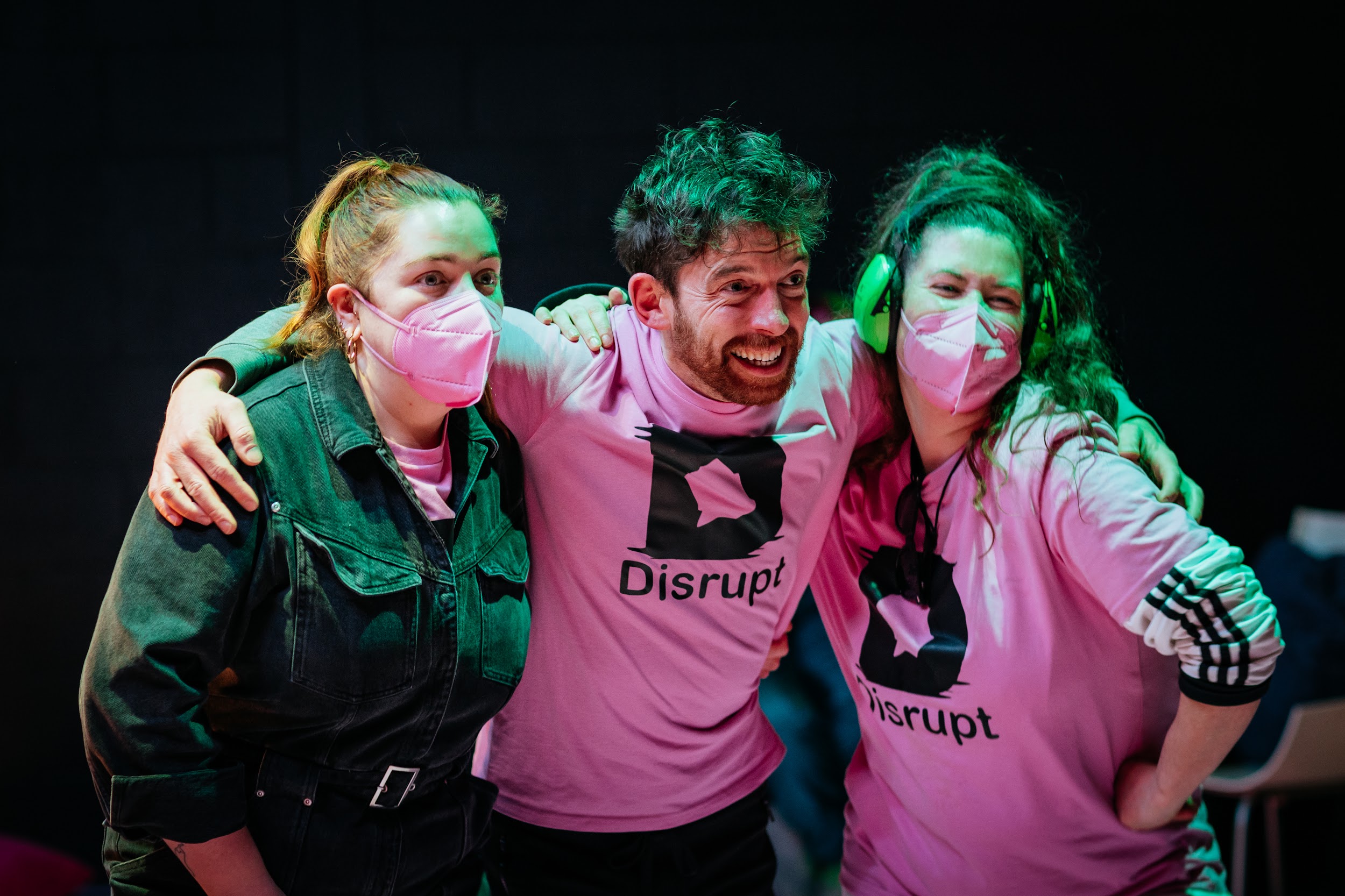 Festival director Alan James Burns poses for a photograph with two Disrupt crew members. They wear pink t-shirts and are lit in green stage lighting. One of them wears ear defenders. All of them wear big smiles.