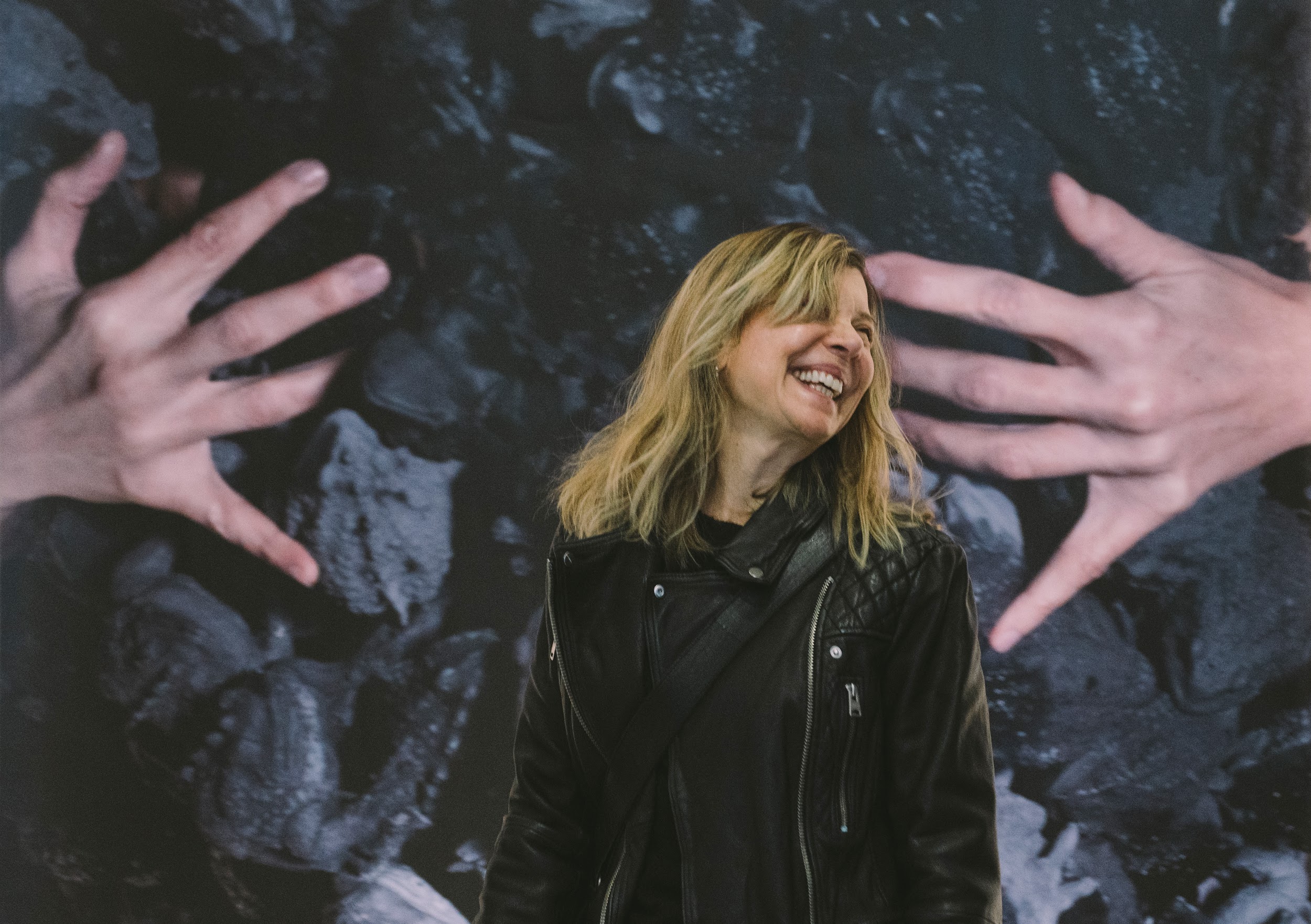 Artist Cecilia Bullo is pictured laughing. She has blonde hair and wears a black biker jacket. They stand in front of a large-scale photographic work, which is partially visible. Photo by Leanne Sullivan.