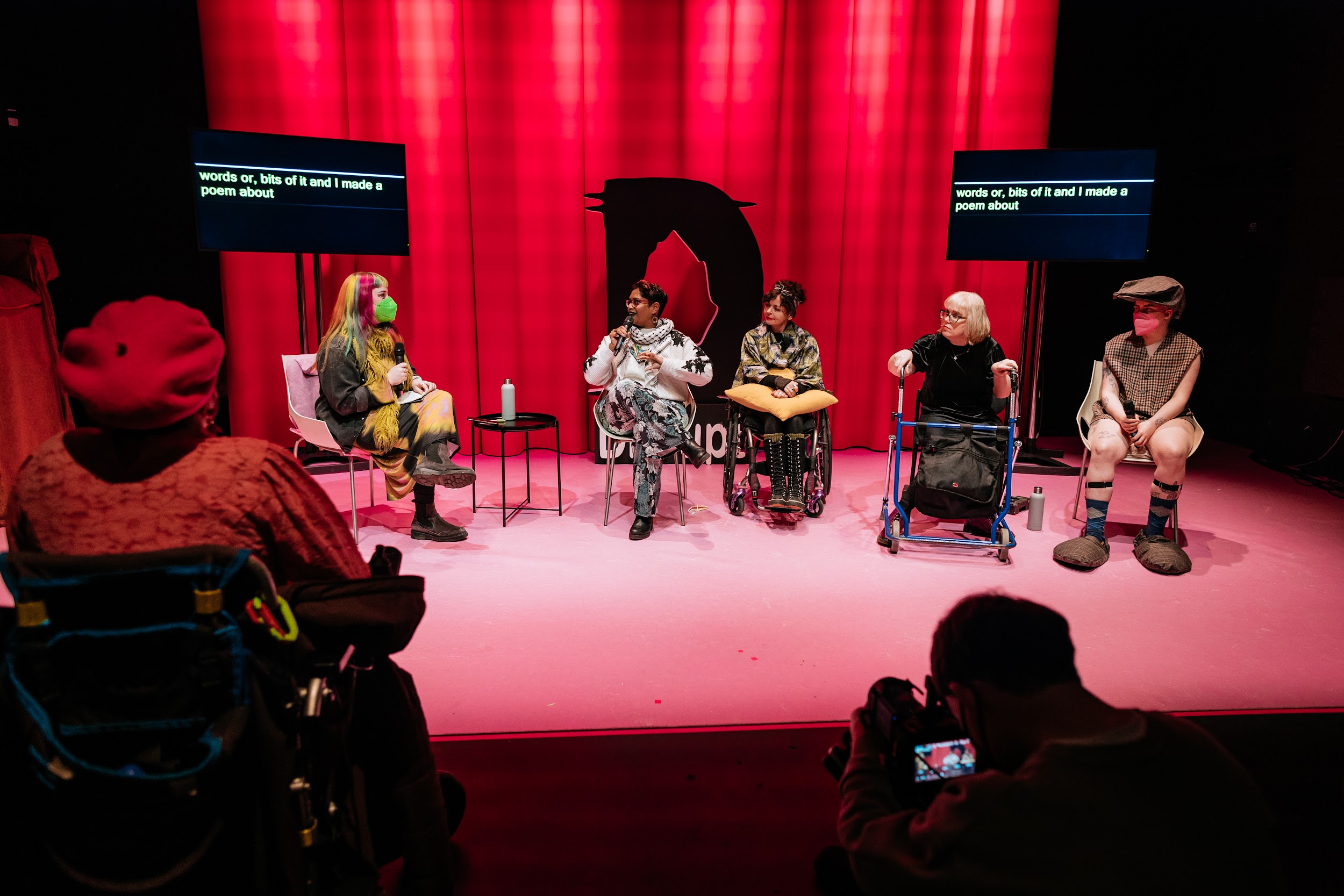 A panel discussion on a stage. Lit in pink. Panellists include Tara Carroll, Chandrika Narayanan-Mohan, Kathy O'Leary, Mairéad Folan, and Them Fatale. Tara holds a mic. Them Fatale is dressed as a caricature of an old man. In the foreground is a person in a red beret who sits in a wheelchair. Photo by Simon Lazewski.