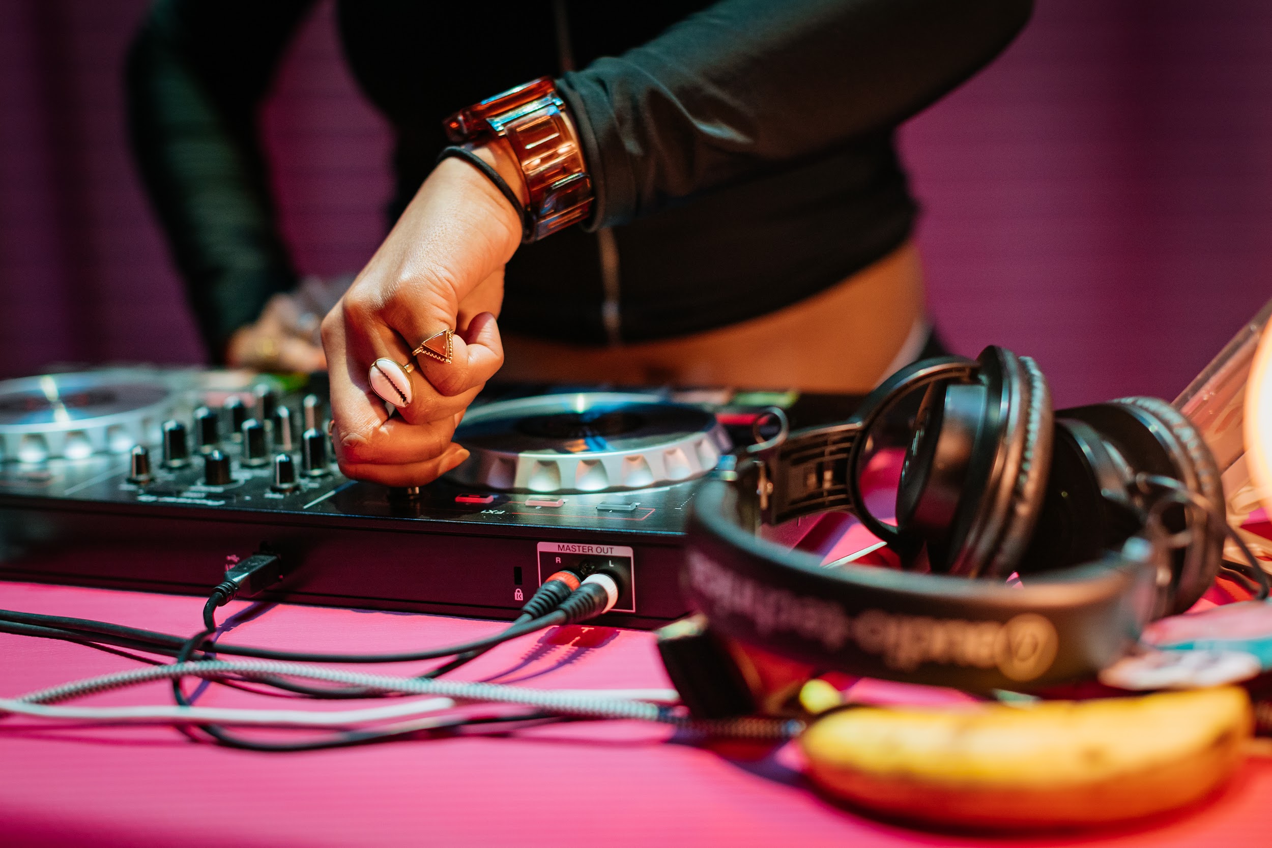 A close up of Dj Renn Miano hands at work. Renn wears black, They have a large tortoiseshell watch and a bobbin on their wrist. They turn a knob on the decks. They wear a shell on a ring and a triangle form on another ring. On the table is a set of large headphones and a banana. Photo by Simon Lazewski.