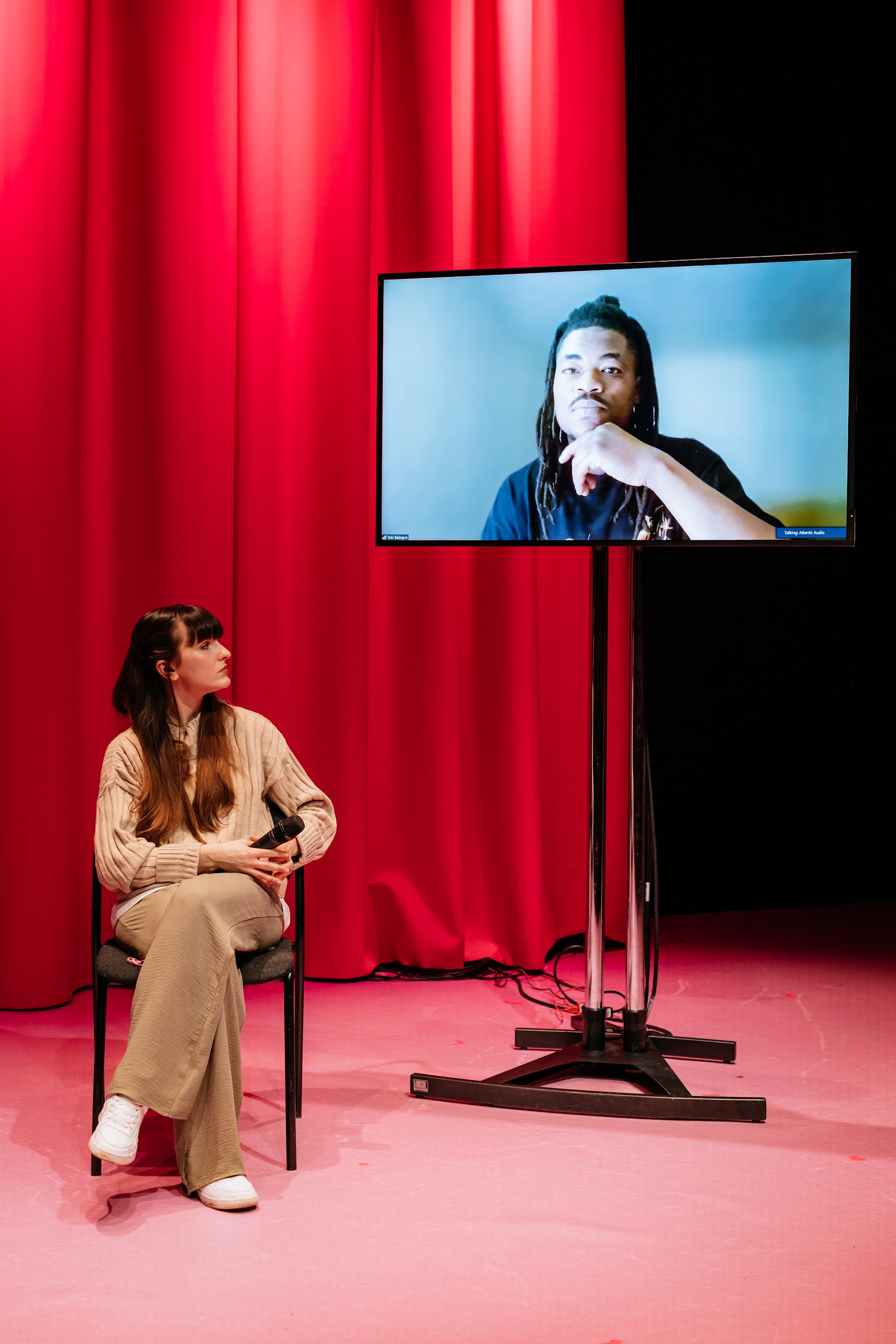 Artist Tobi Balogun is seen through a videolink, on a monitor. Via videolink he is onstage. Behind the screen hangs a pink theatre curtain. Tobi is captured from the shoulders up. He is pensive, about to speak. Dancer Alison Clarke, on stage in person, looks on. Photo by Simon Lazewski.