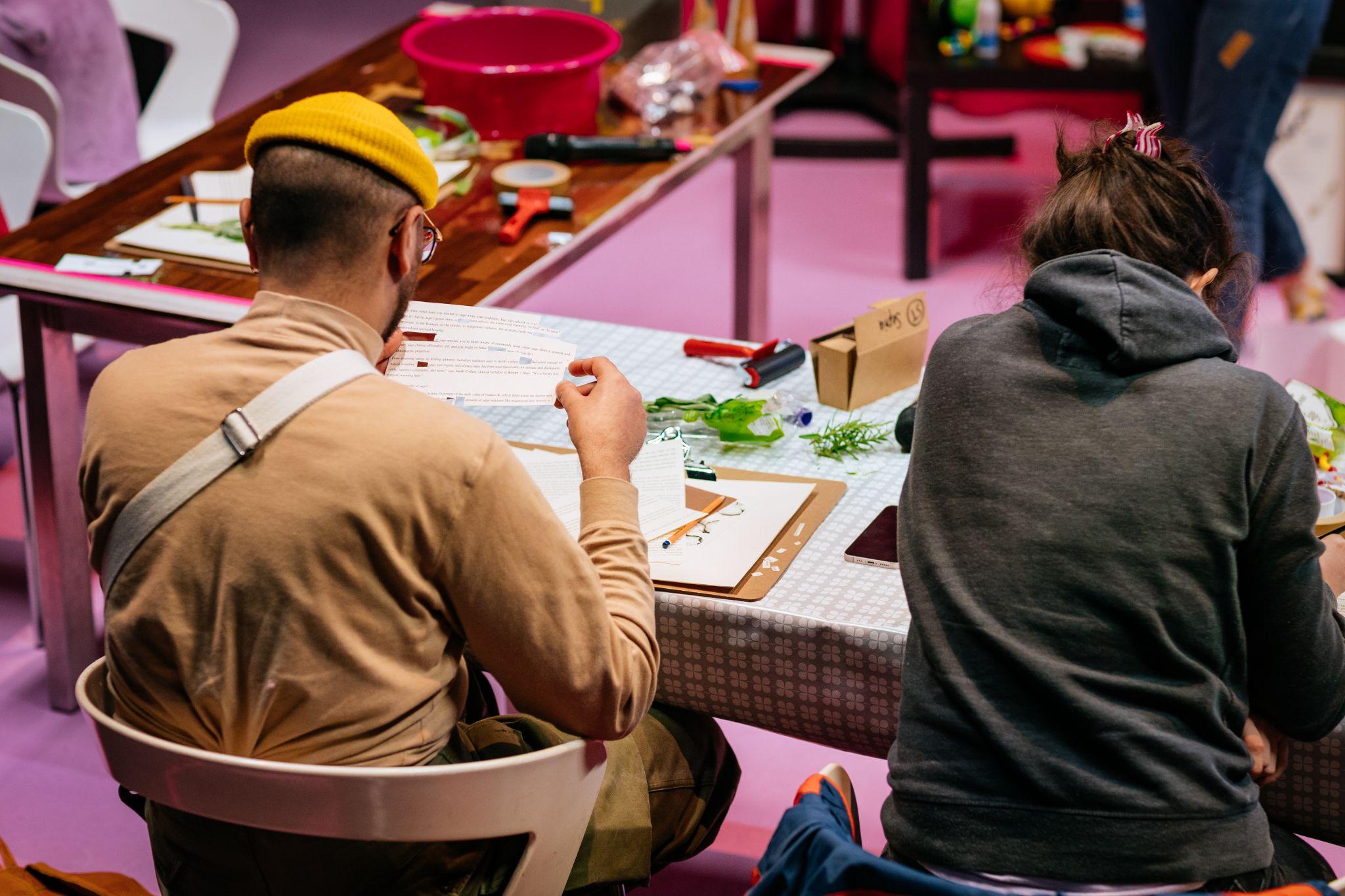 An image taken from behind shows two people sitting at a desk. The person on the left has short dark hair and dark skin, wearing glasses, a yellow hat, and a light brown polo neck top. They appear to be cutting words from a page of text with scissors. The person on the right wears a dark-coloured hooded top and has long dark hair. Various materials, including more texts, small boxes, and paints, are on the table in front of them. In the blurry background are other desks with materials. Photo by Simon Lazewski.