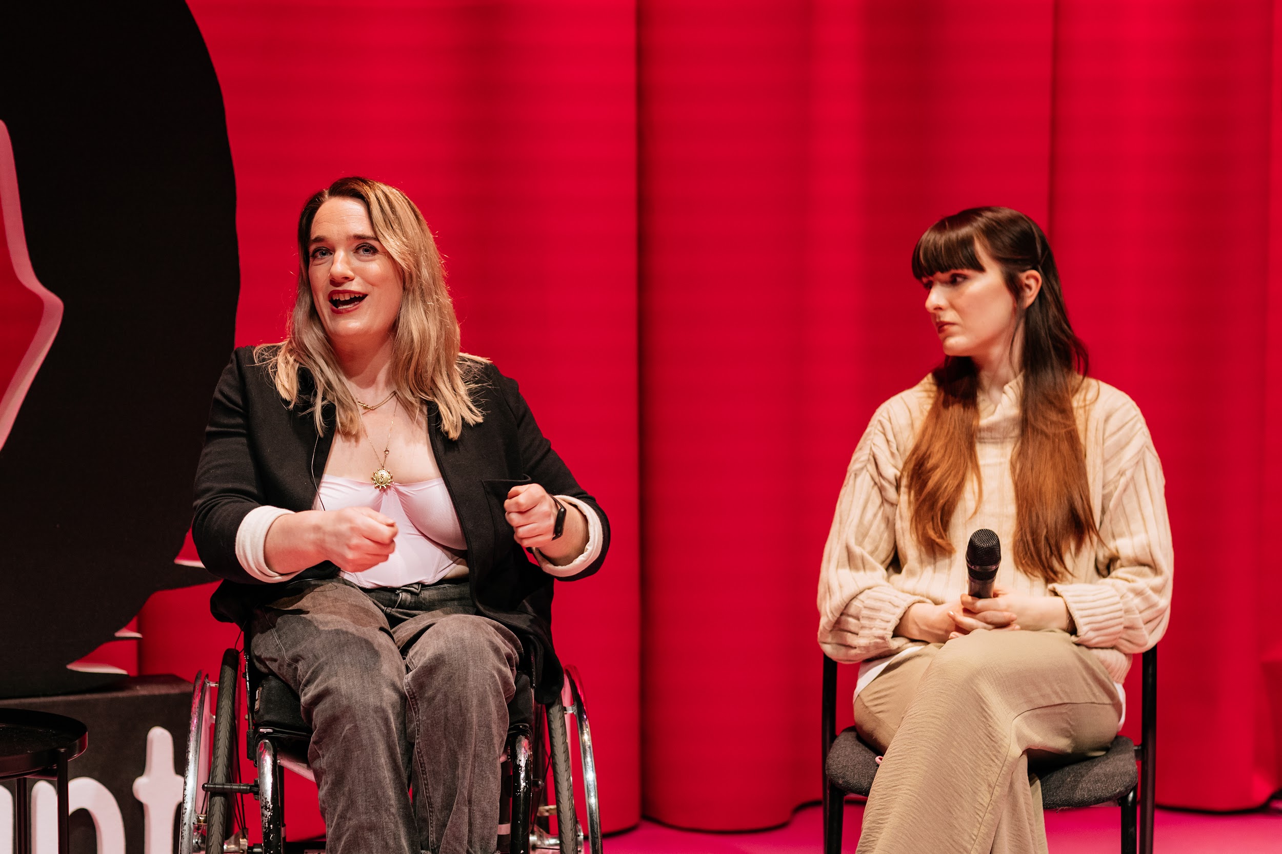 On the Disrupt Disability Arts stage, sit two individuals. They are Louise Bruton and Alison Clarke. They are engaged in conversation with somebody who is out of shot. Louise, a blond woman who is a wheelchair user, is speaking in an animated fashion and gesticulates with her hands. To the right sits Alison, who looks on. Alison wears camel colours and holds a microphone on her lap.